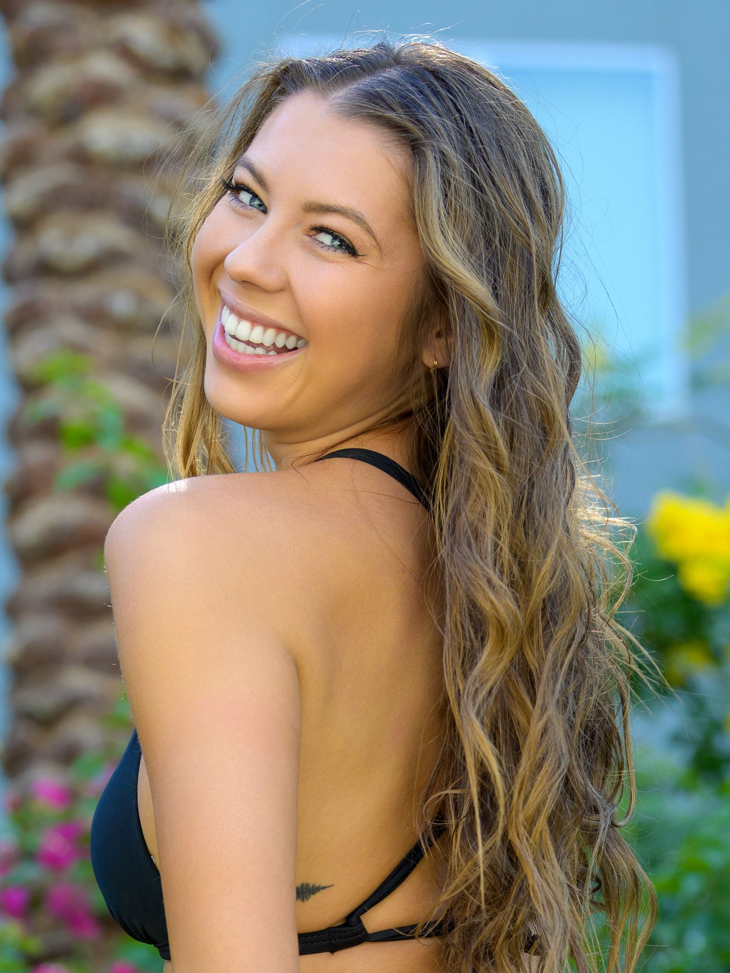 Smiling young woman with long wavy blonde hair wearing a black bikini top, posing outdoors near tropical plants.