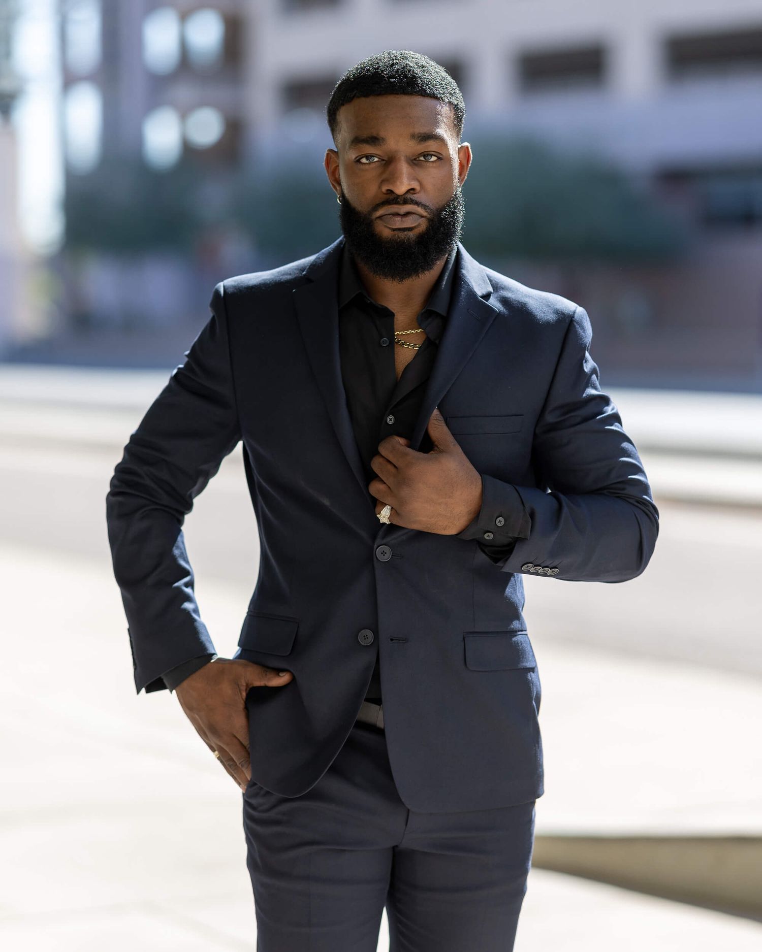 A confident Black man in a navy blue suit and black shirt poses outdoors in an urban setting.