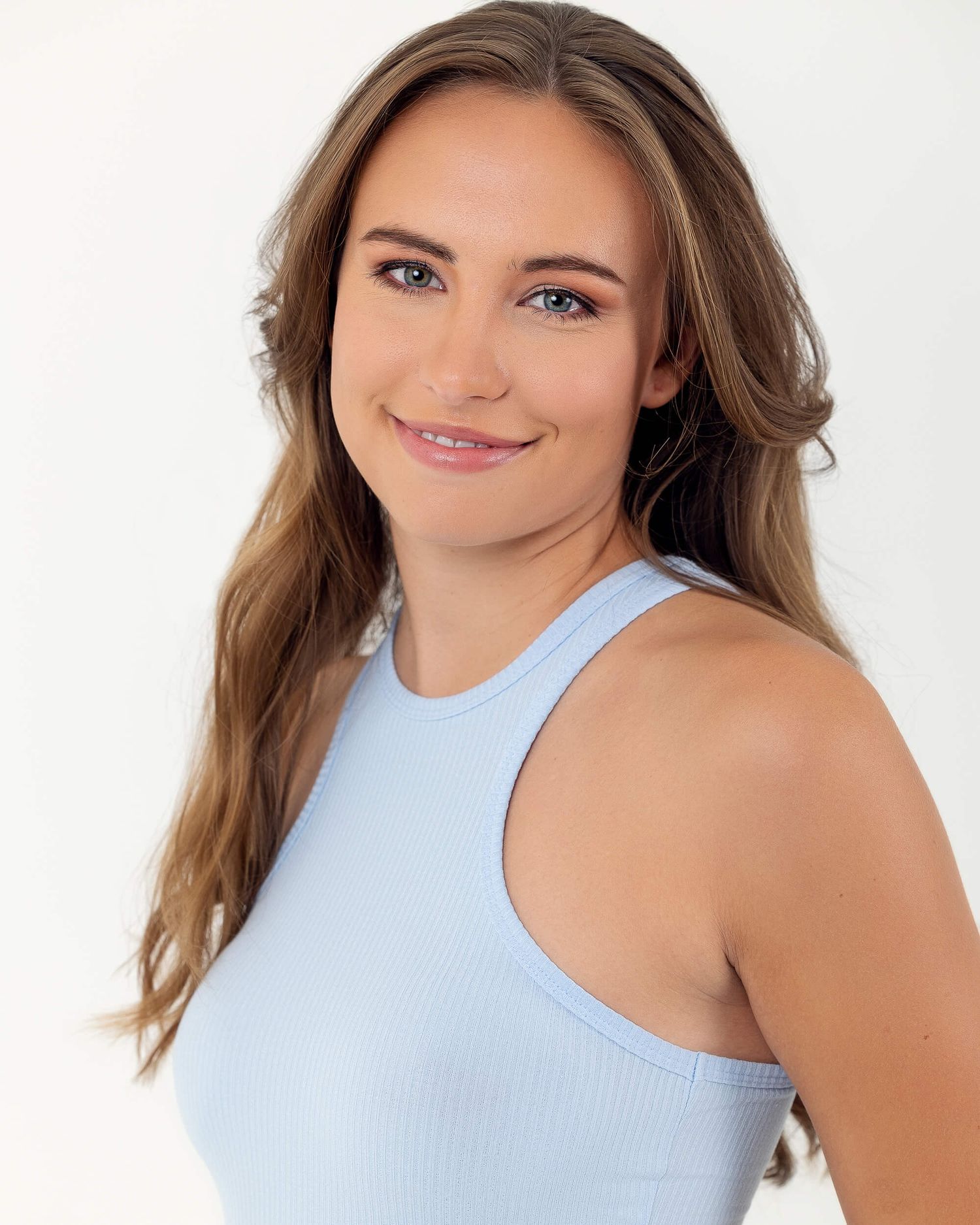 Young woman with long brown hair smiling in a light blue tank top against white background.