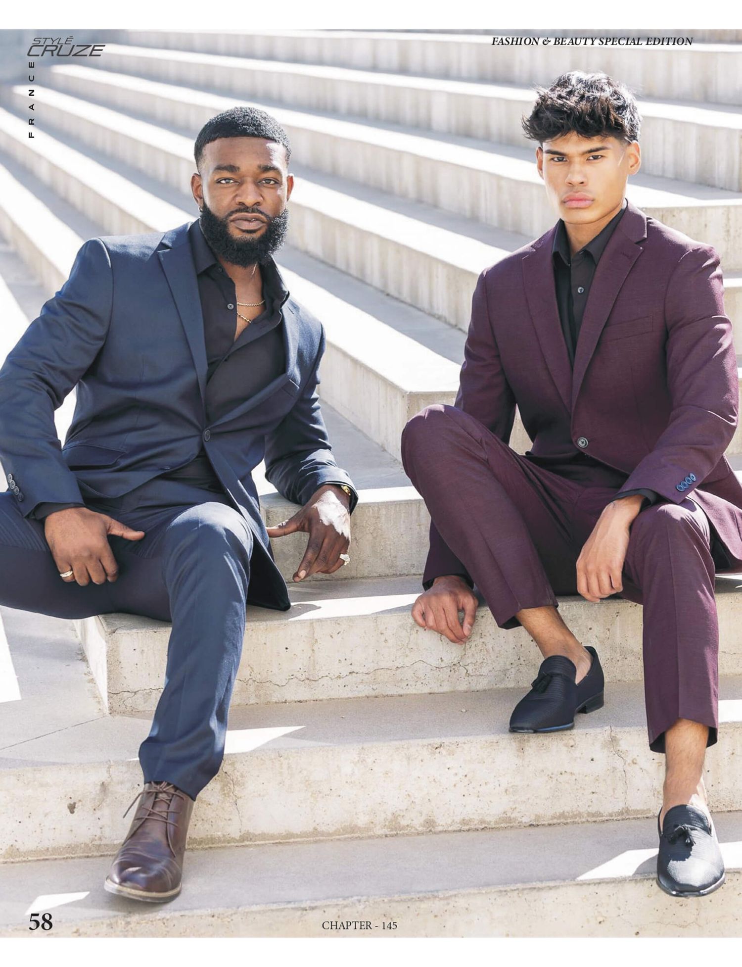 Two stylish men in navy and burgundy suits sit on outdoor steps in a fashion and beauty special edition spread.
