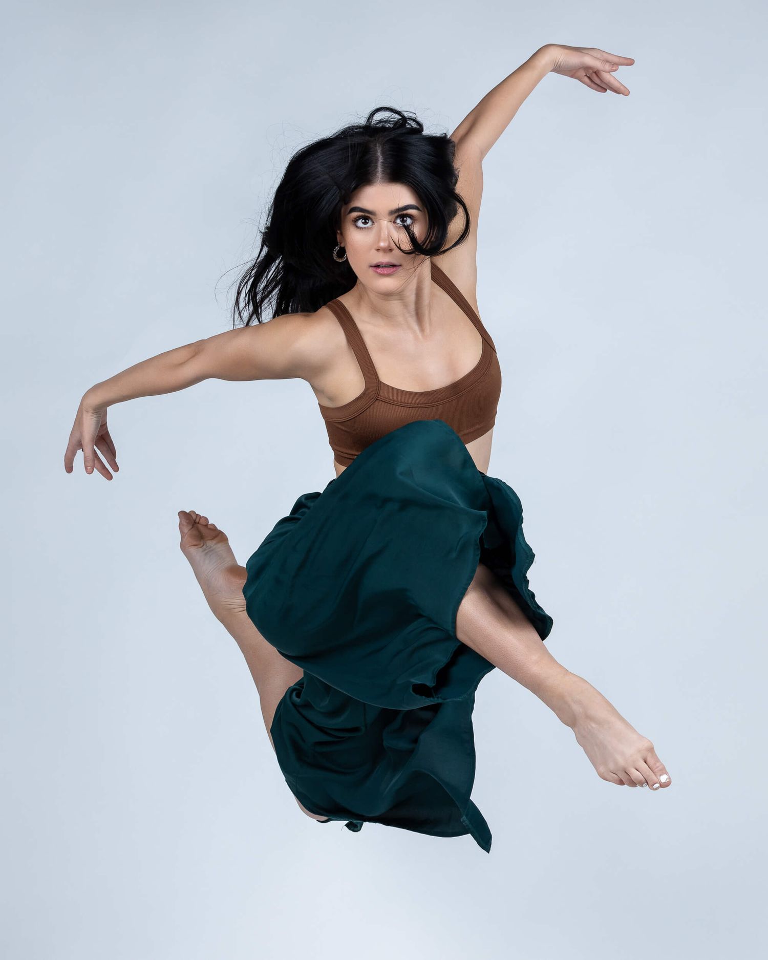 Dynamic female dancer leaps gracefully in a brown crop top and flowing teal skirt against a light backdrop.