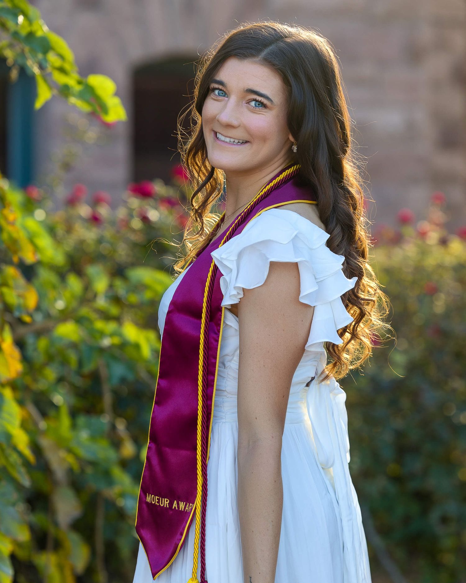 Smiling graduate in white dress wearing maroon honor stole posing in front of lush garden greenery.