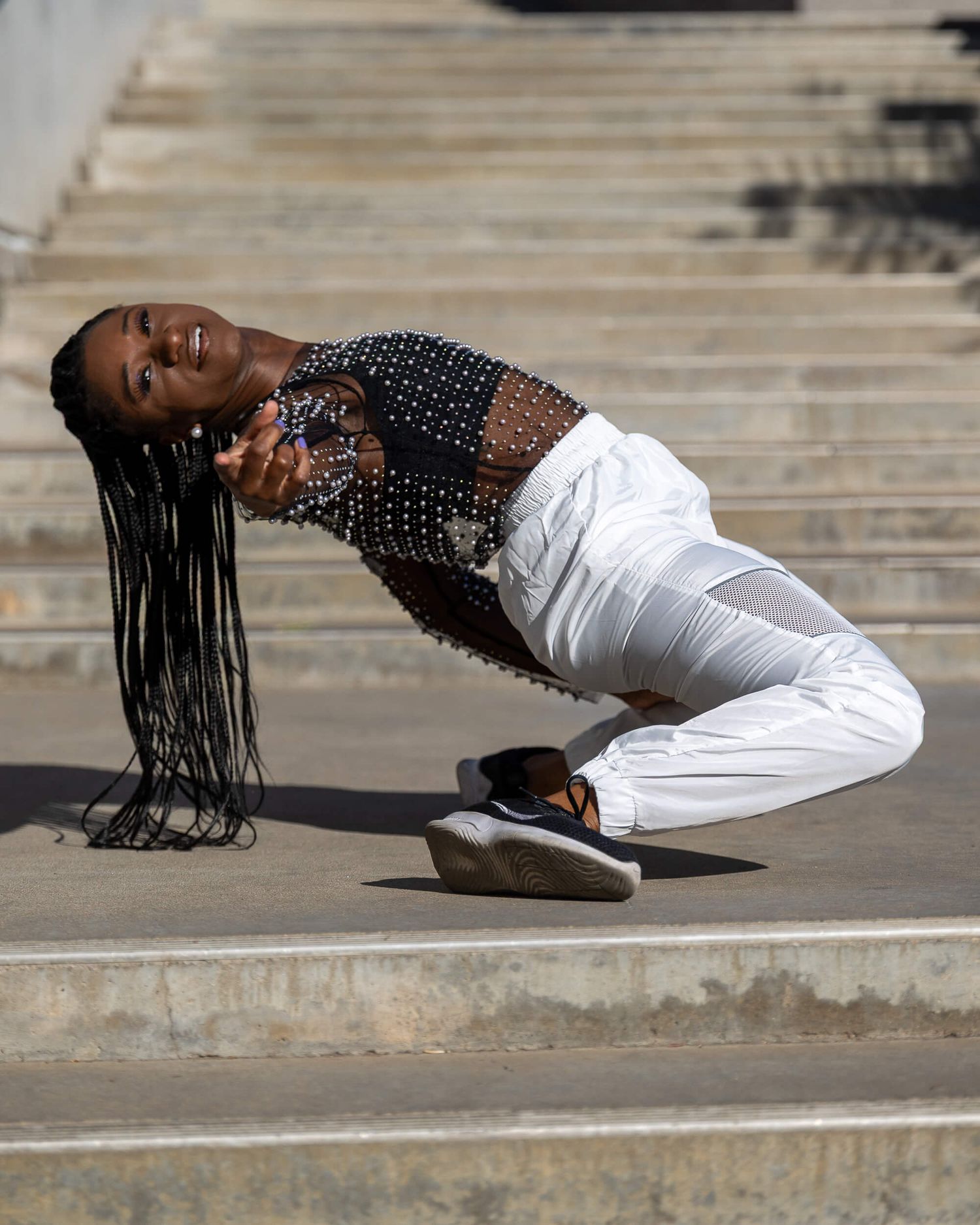 Female dancer in white pants and studded top performs low backbend on outdoor steps.