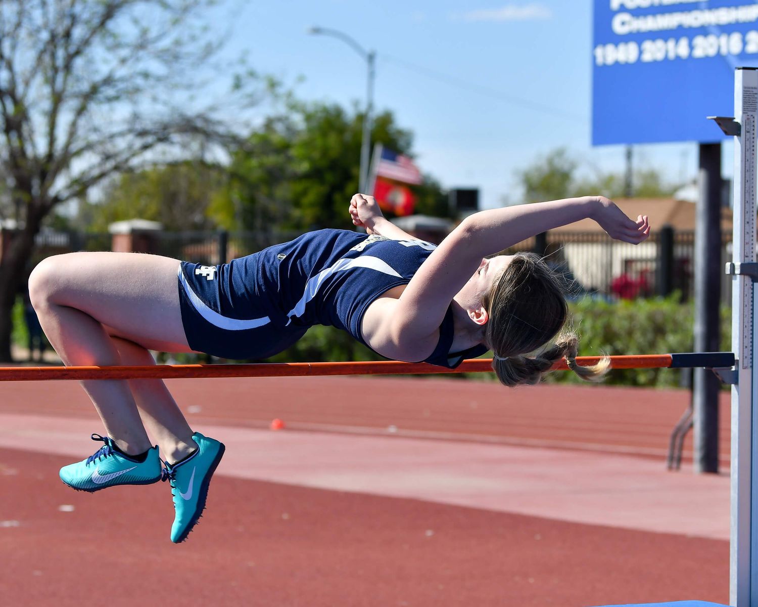 Female high jumper in navy uniform performs the Fosbury Flop technique over the bar at a track meet.