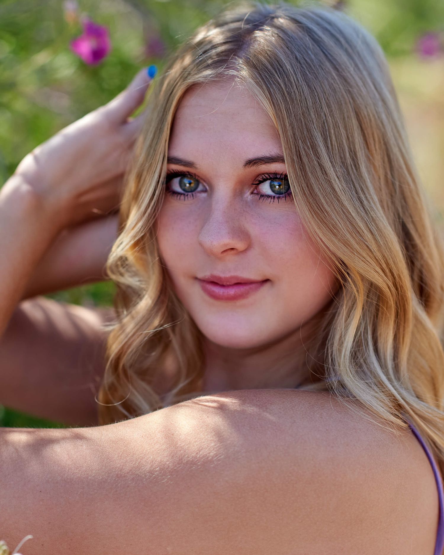 Young blonde woman with blue eyes and freckles poses outdoors near pink flowers.