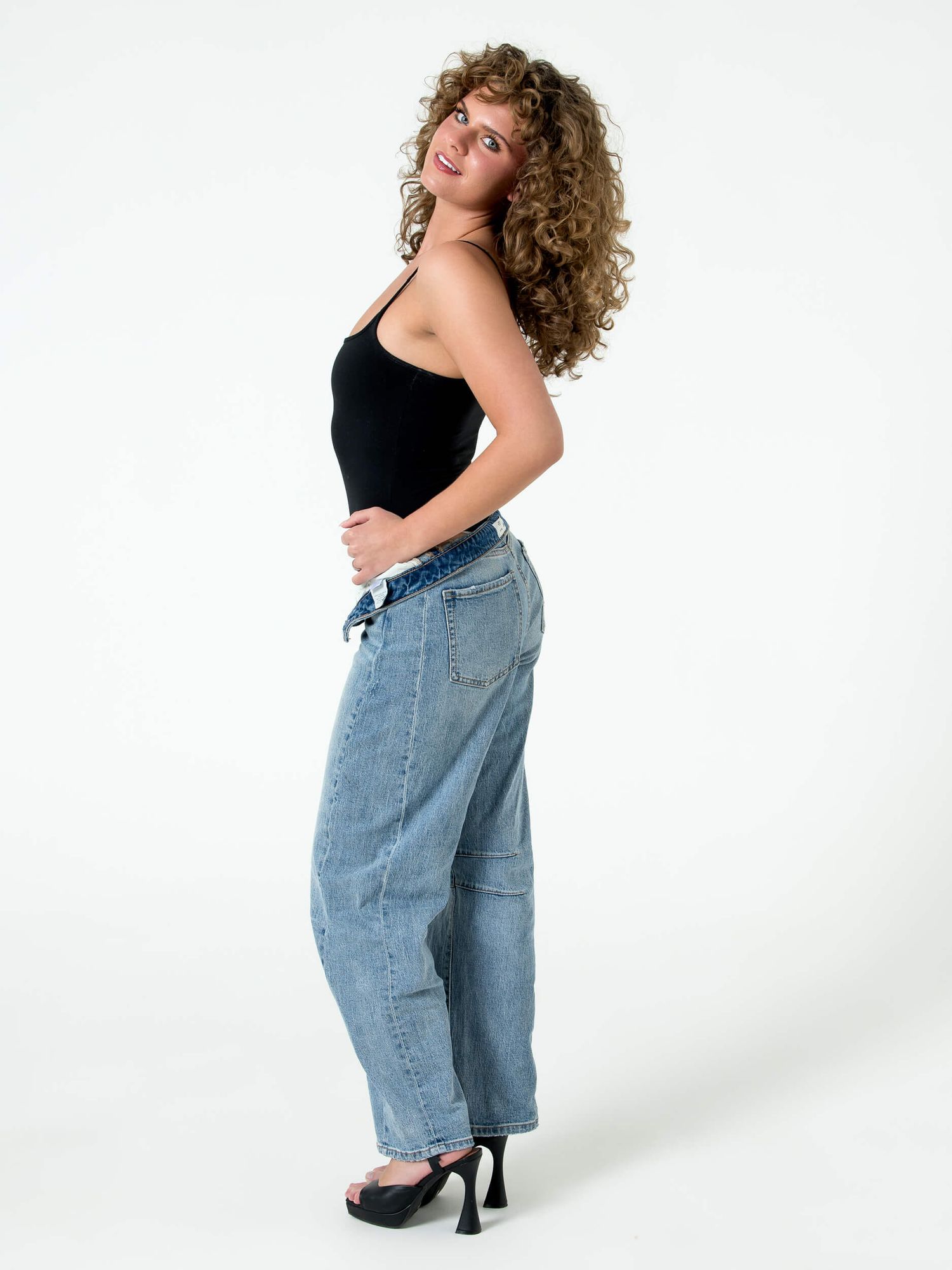 Woman in black tank top and light wash baggy jeans poses sideways in black heels against white background.
