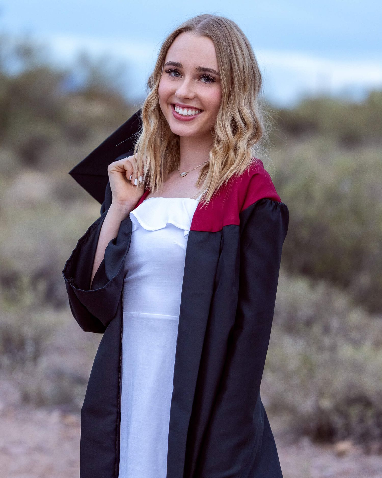 Blonde woman in black and burgundy graduation gown smiling outdoors in desert landscape.