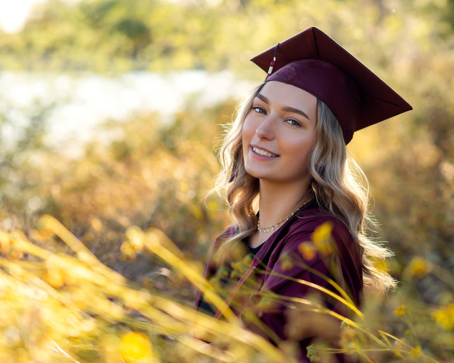 Young blonde woman smiling in maroon graduation cap and gown outdoors among golden wildflowers.