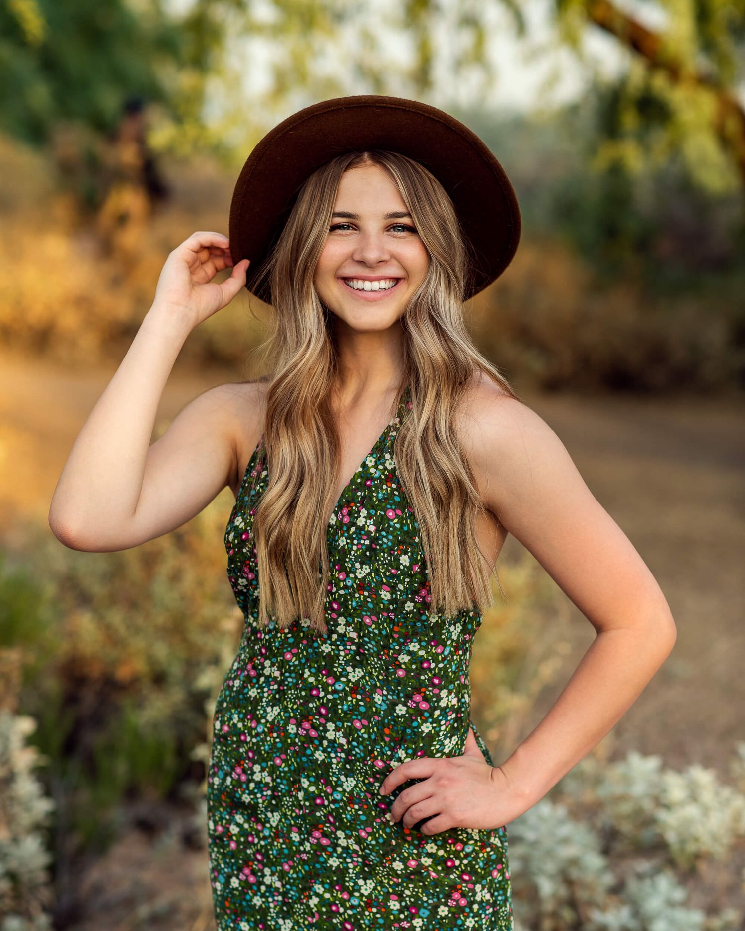 Smiling young woman in floral green dress and brown hat posing outdoors in golden light.