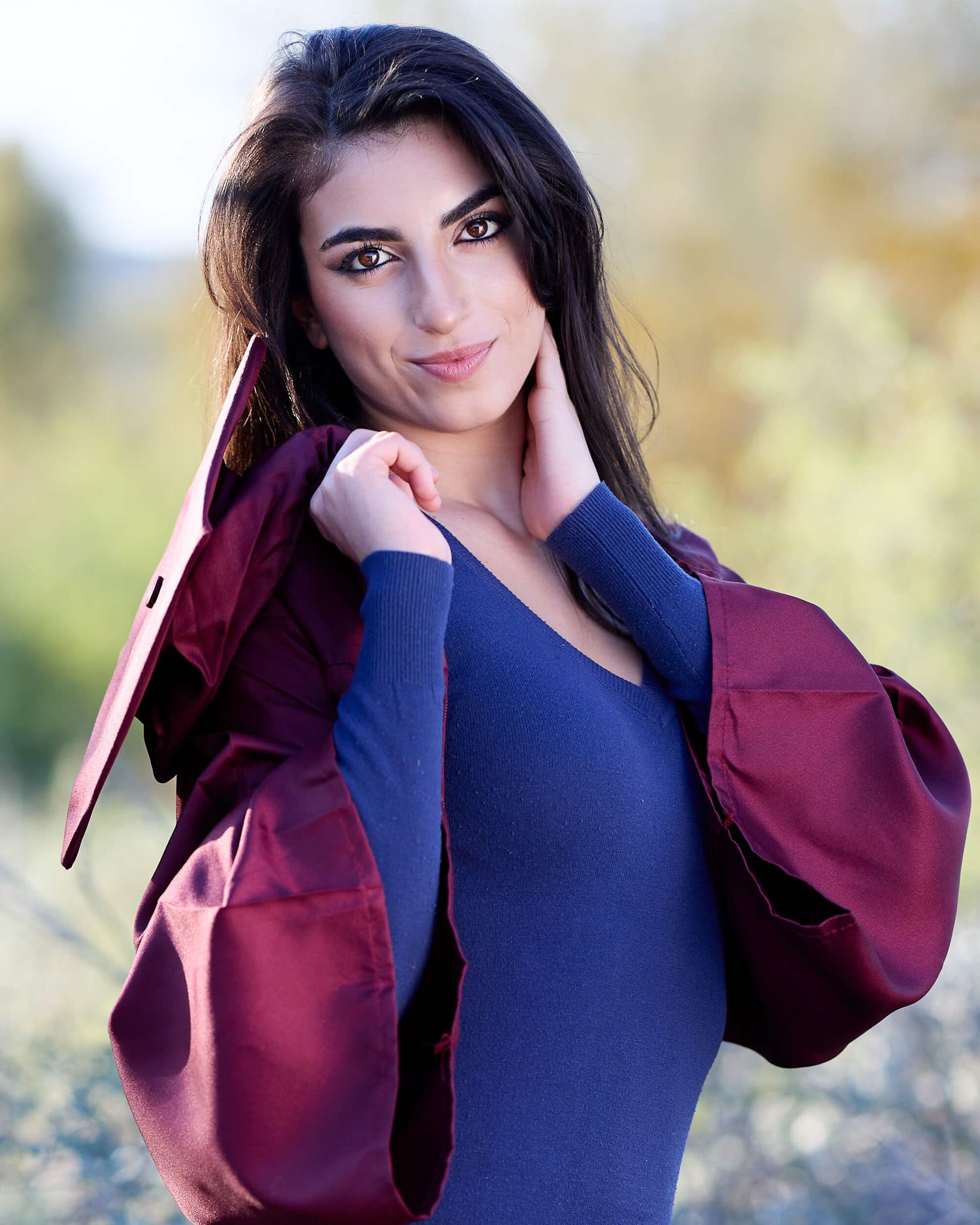 Young woman in blue dress holding burgundy graduation gown over shoulder, smiling outdoors.
