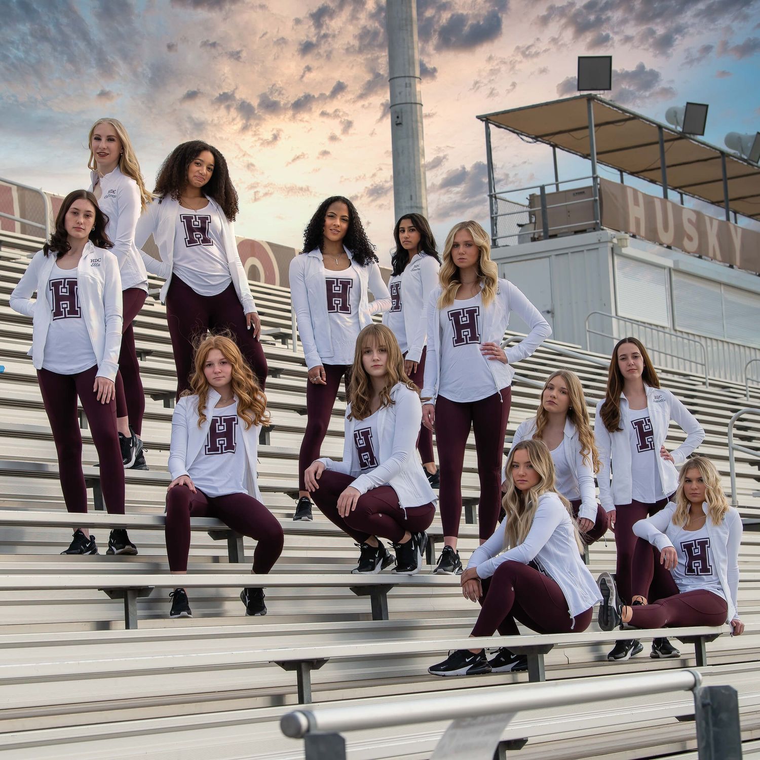 Dance team posing in white and maroon uniforms on stadium bleachers at sunset.