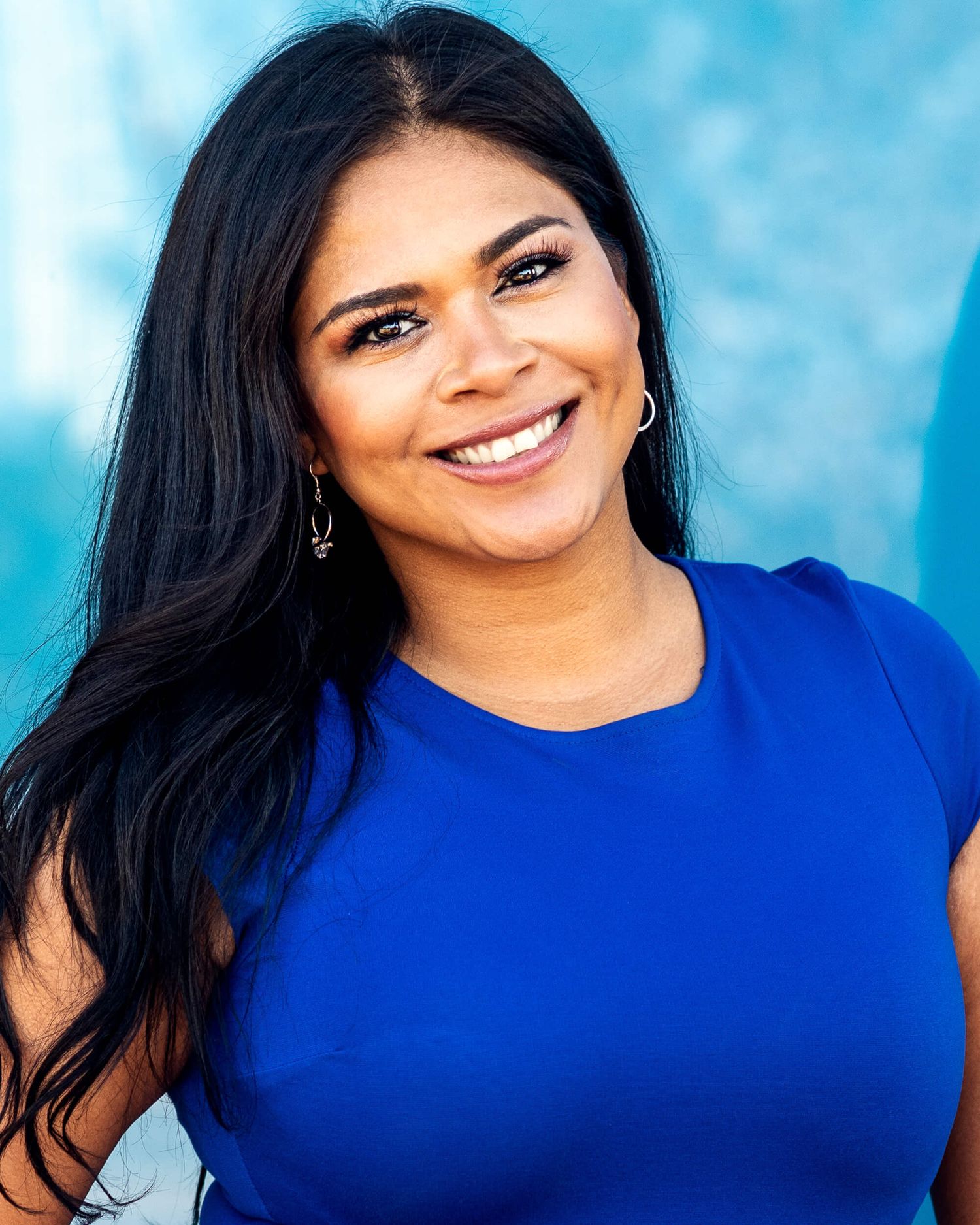 Smiling woman with long black hair wearing royal blue top against turquoise background.