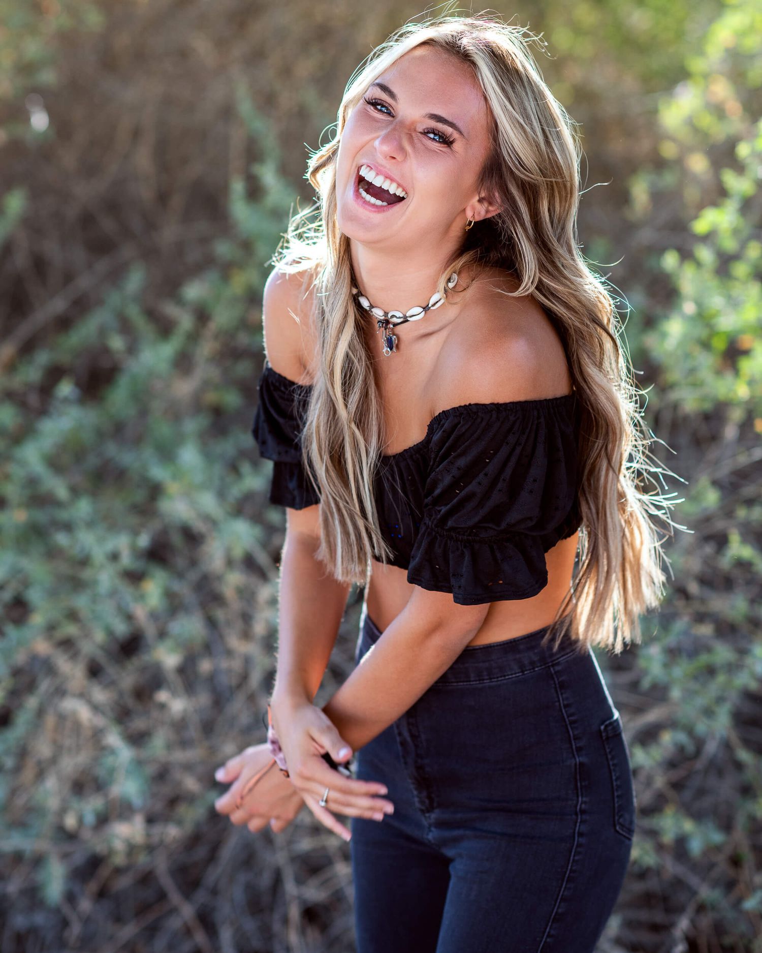 Laughing blonde woman in black off-shoulder crop top and jeans posing in natural setting.