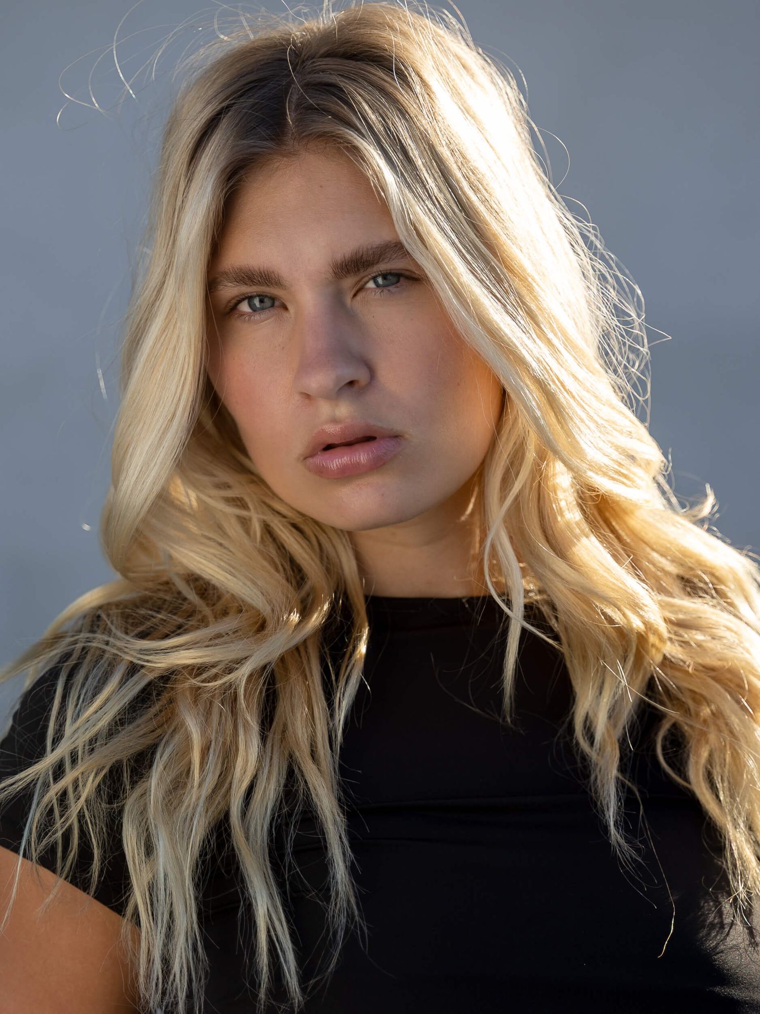 Young woman with long blonde wavy hair wearing a black top, posing outdoors with a confident expression.