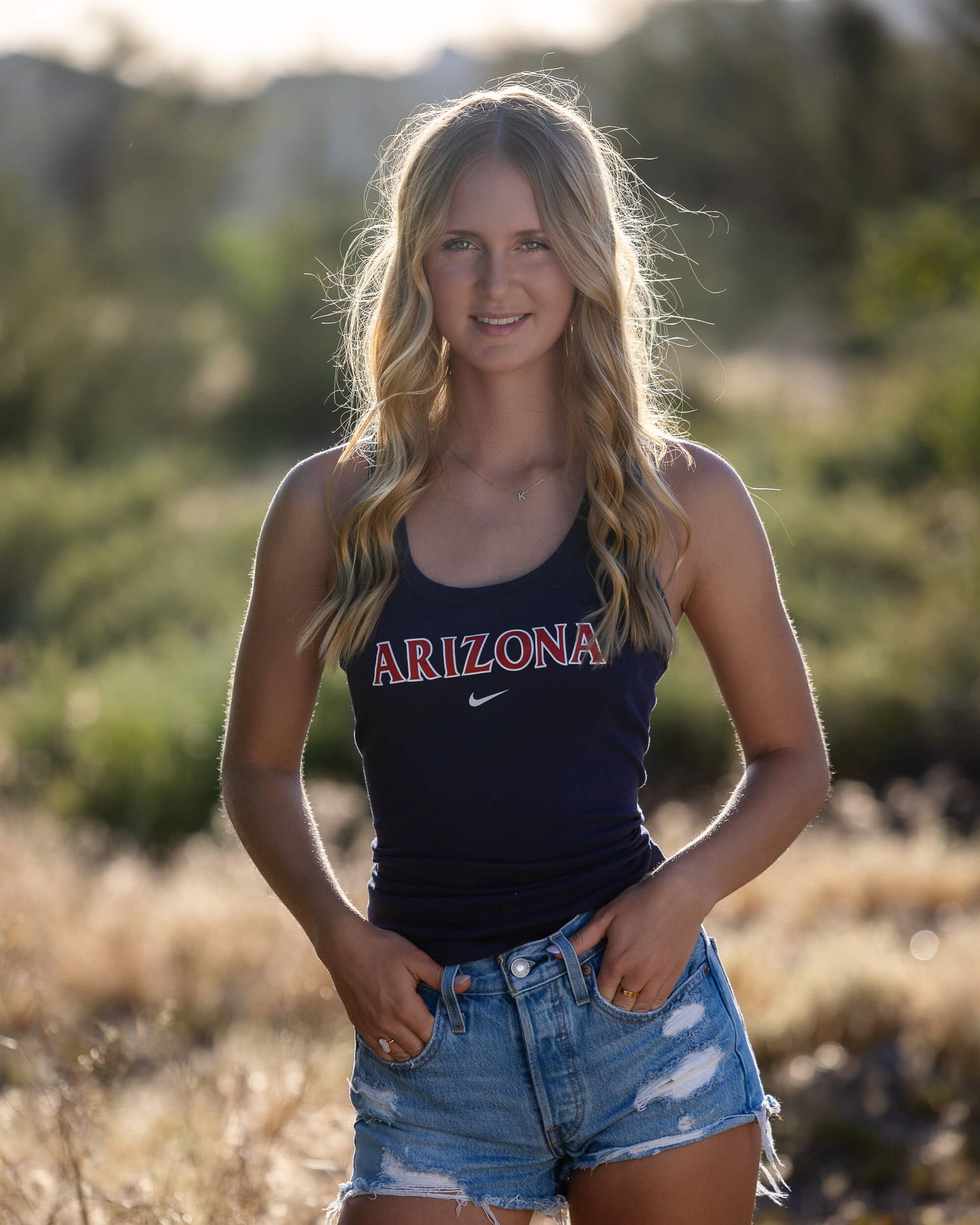 Young woman in Arizona Nike tank top and denim shorts smiling outdoors in a sunny natural desert landscape.