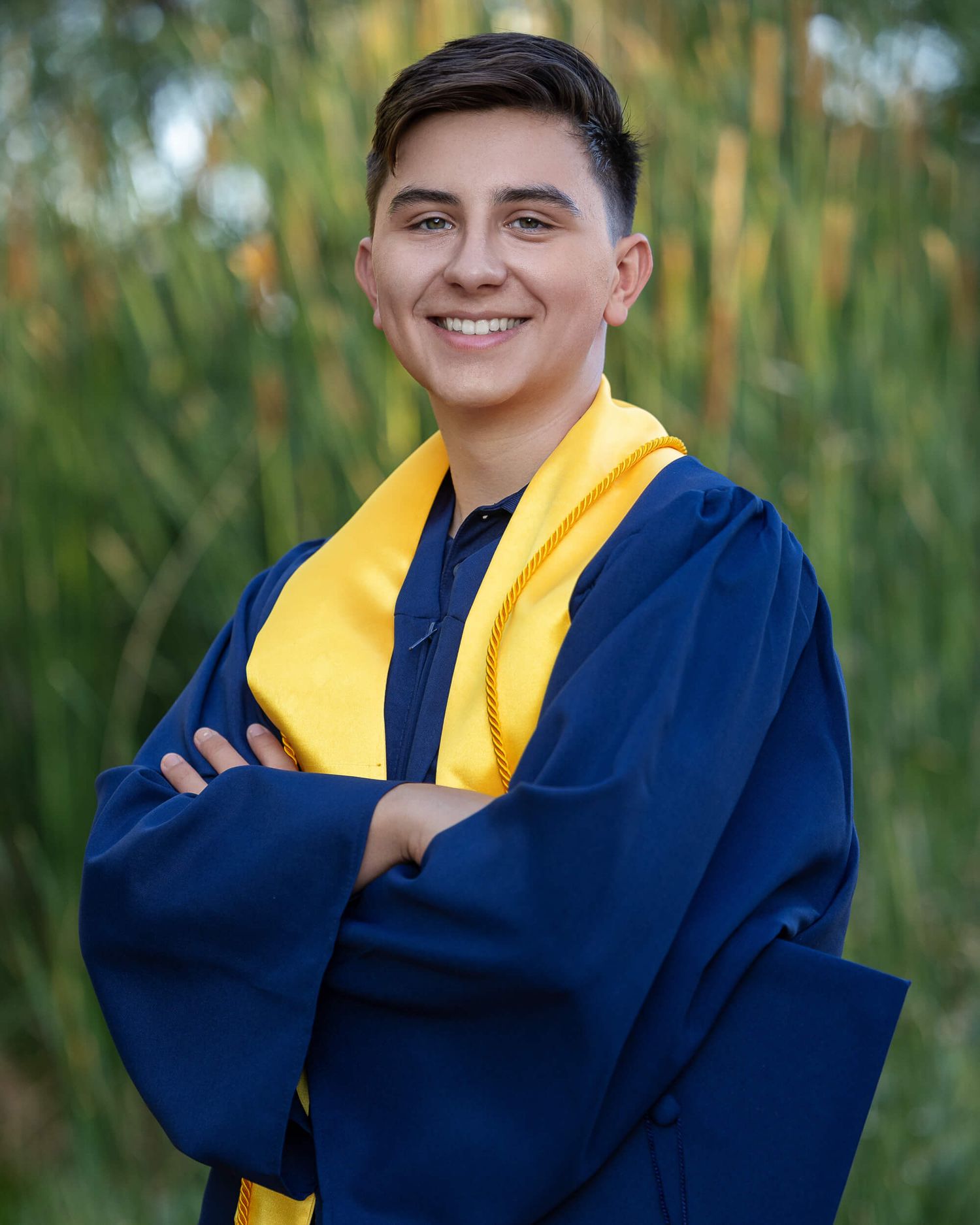 Smiling graduate in navy blue cap and gown with gold sash, arms crossed, posing outdoors.