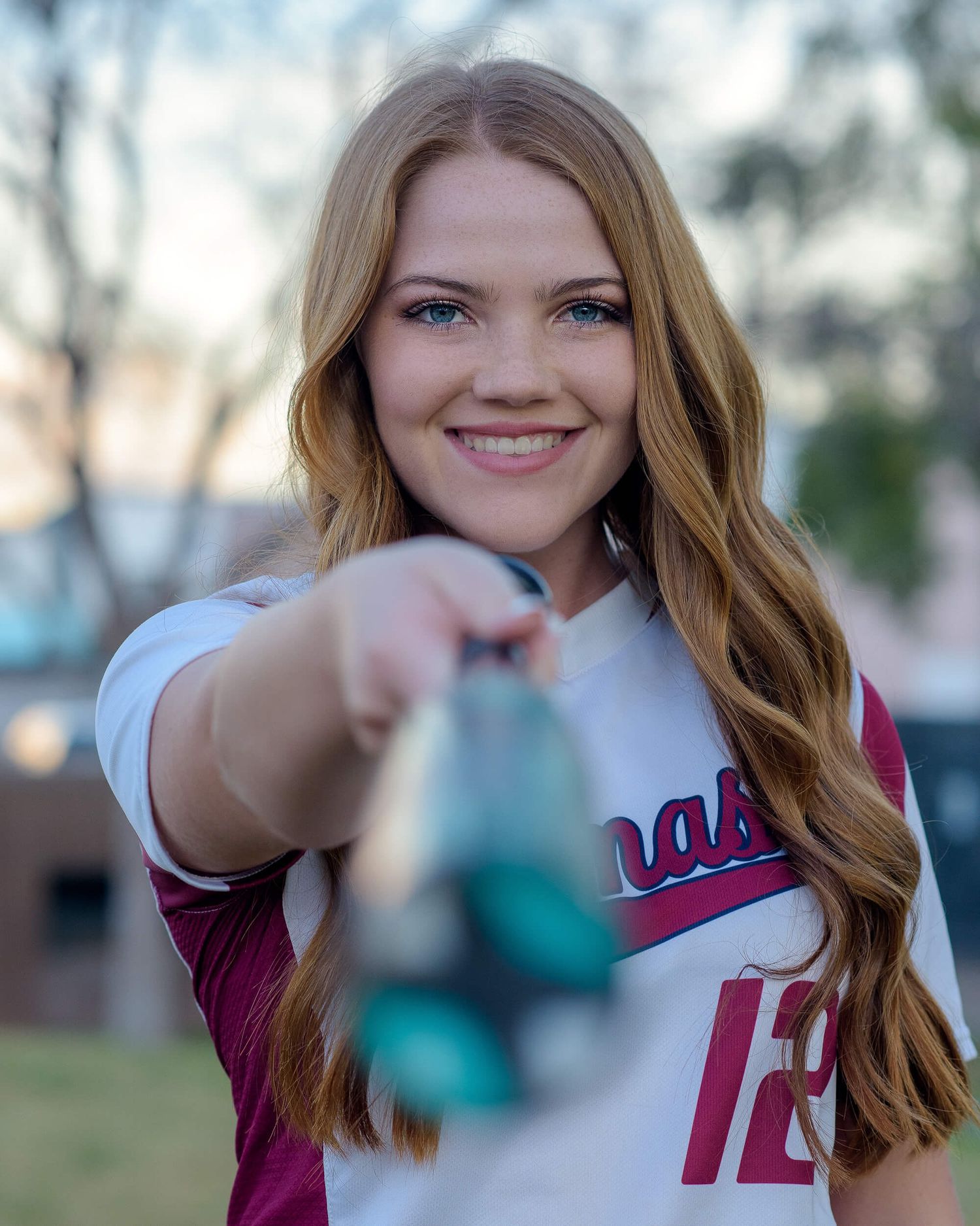 Smiling young woman in softball jersey holding ball toward camera outdoors.