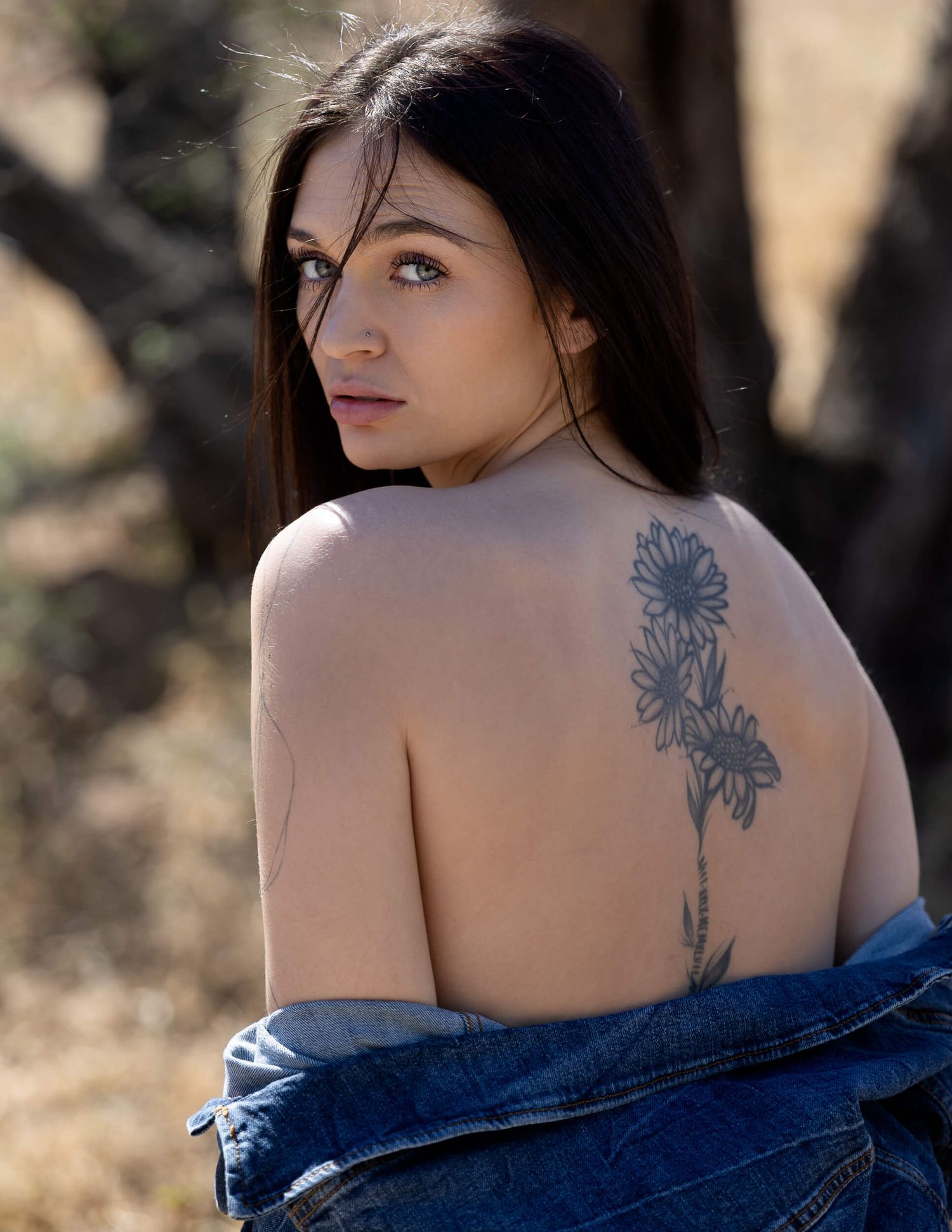 Young woman with dark hair and floral back tattoo posing outdoors in a denim jacket against a natural background.
