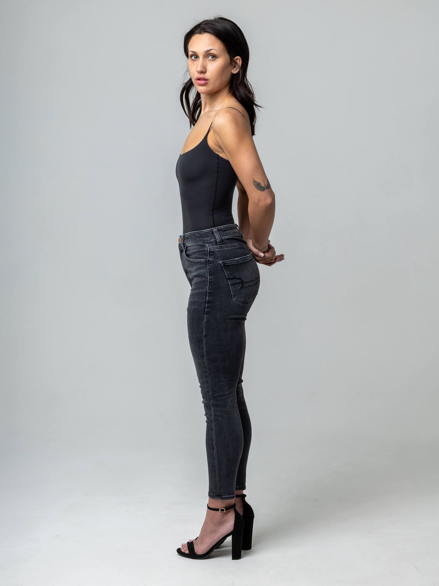 Woman in black bodysuit and dark skinny jeans with heeled sandals, posing sideways with a tattoo on her arm.