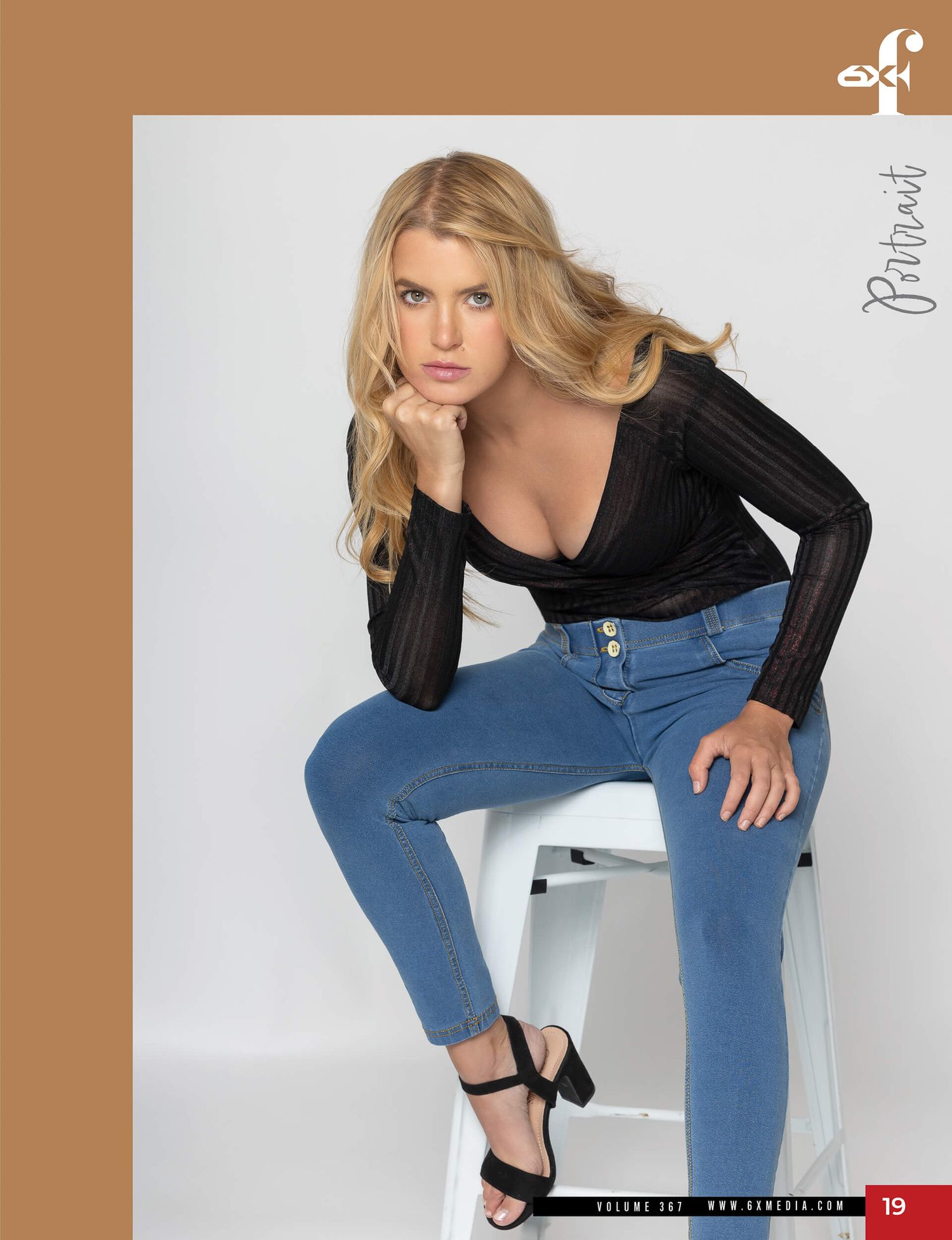 Blonde woman in deep V-neck black top and blue jeans seated on stool.
