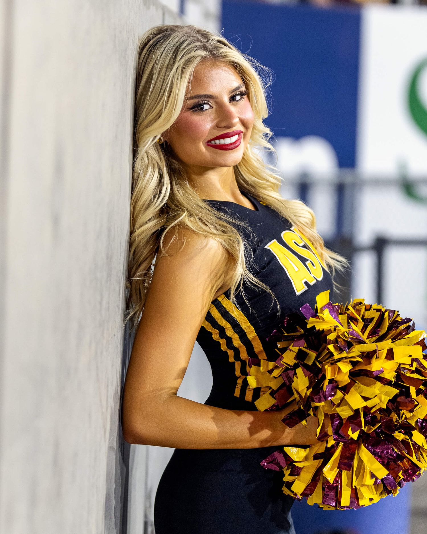 ASU cheerleader in black uniform poses with gold pom-poms against a wall.