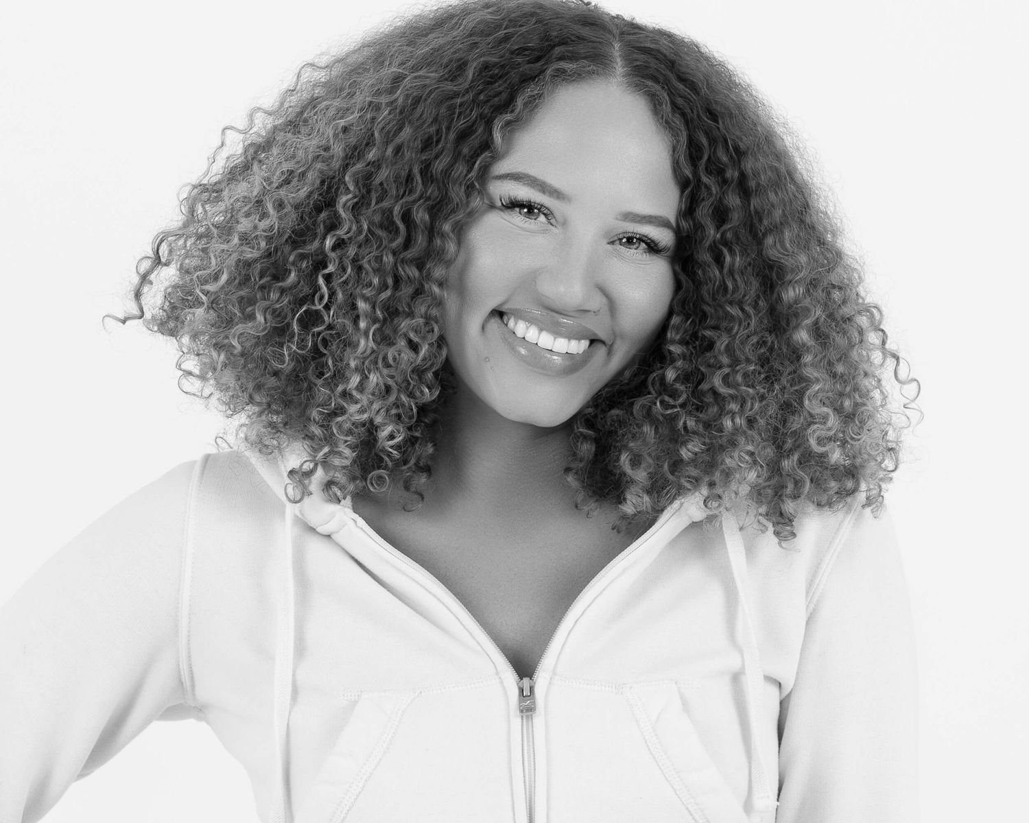 Black and white photo of a smiling young woman with voluminous curly hair in a white hoodie.