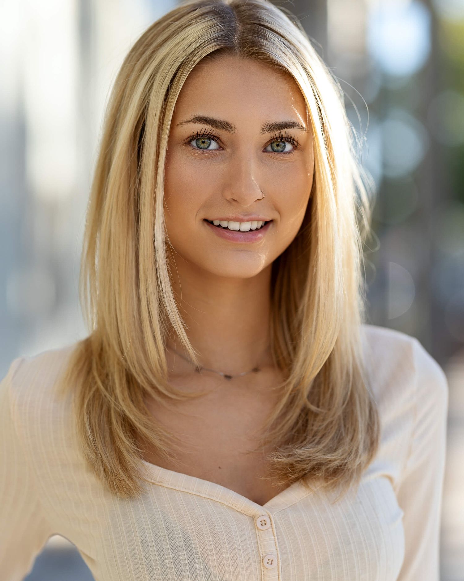 Young woman with long blonde hair and green eyes smiling in a white top outdoors.