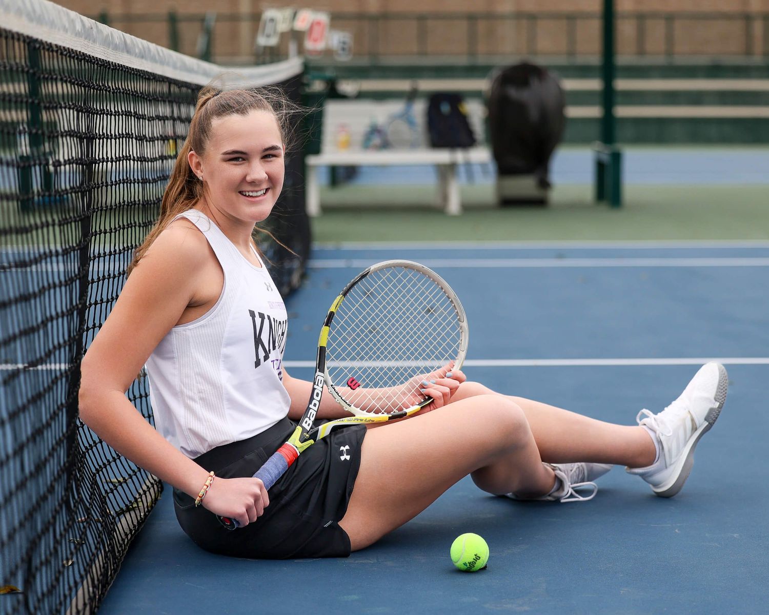 Smiling female tennis player sits courtside holding racket with tennis ball nearby.