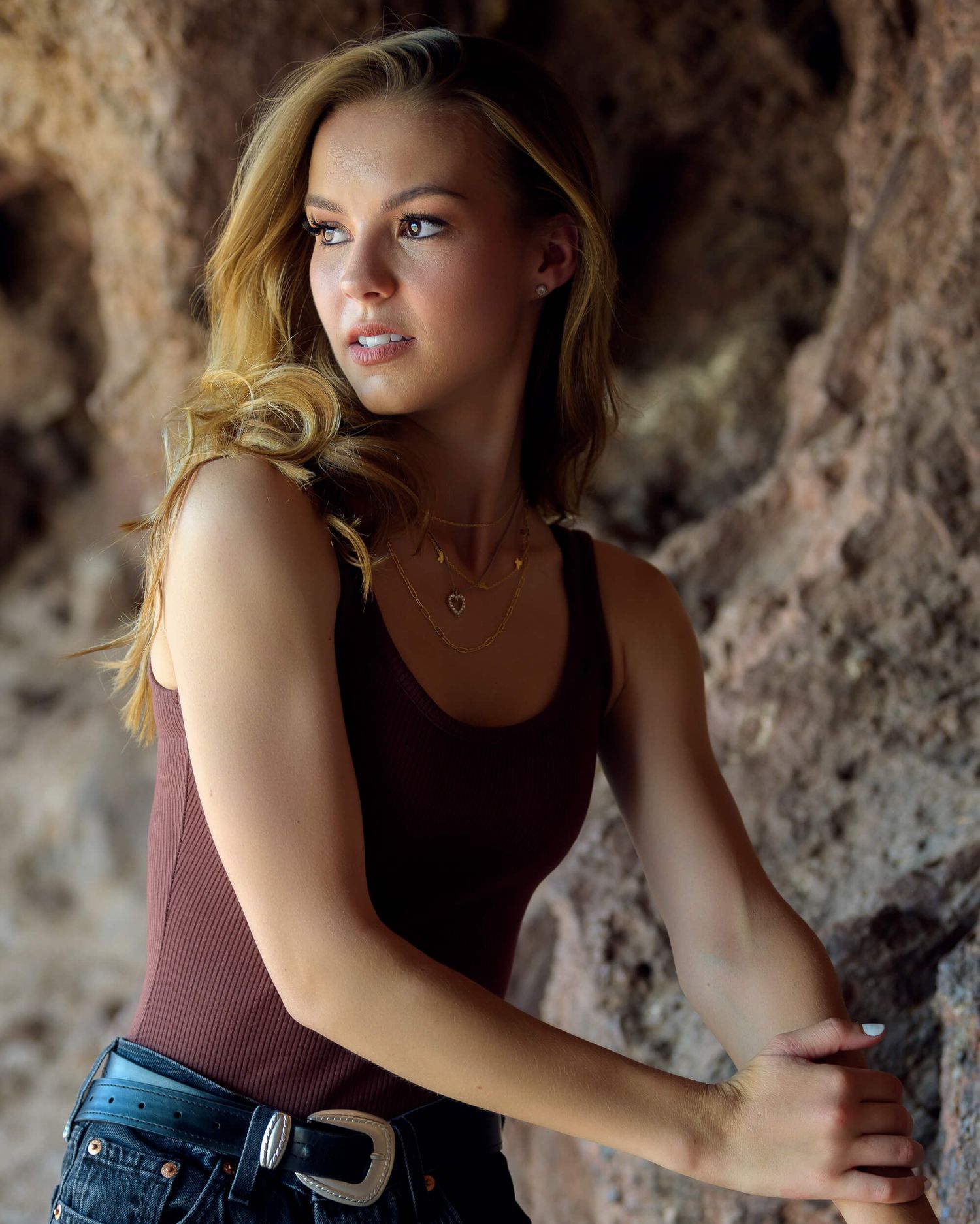Blonde woman in brown tank top and jeans posing against rocky cave wall background.
