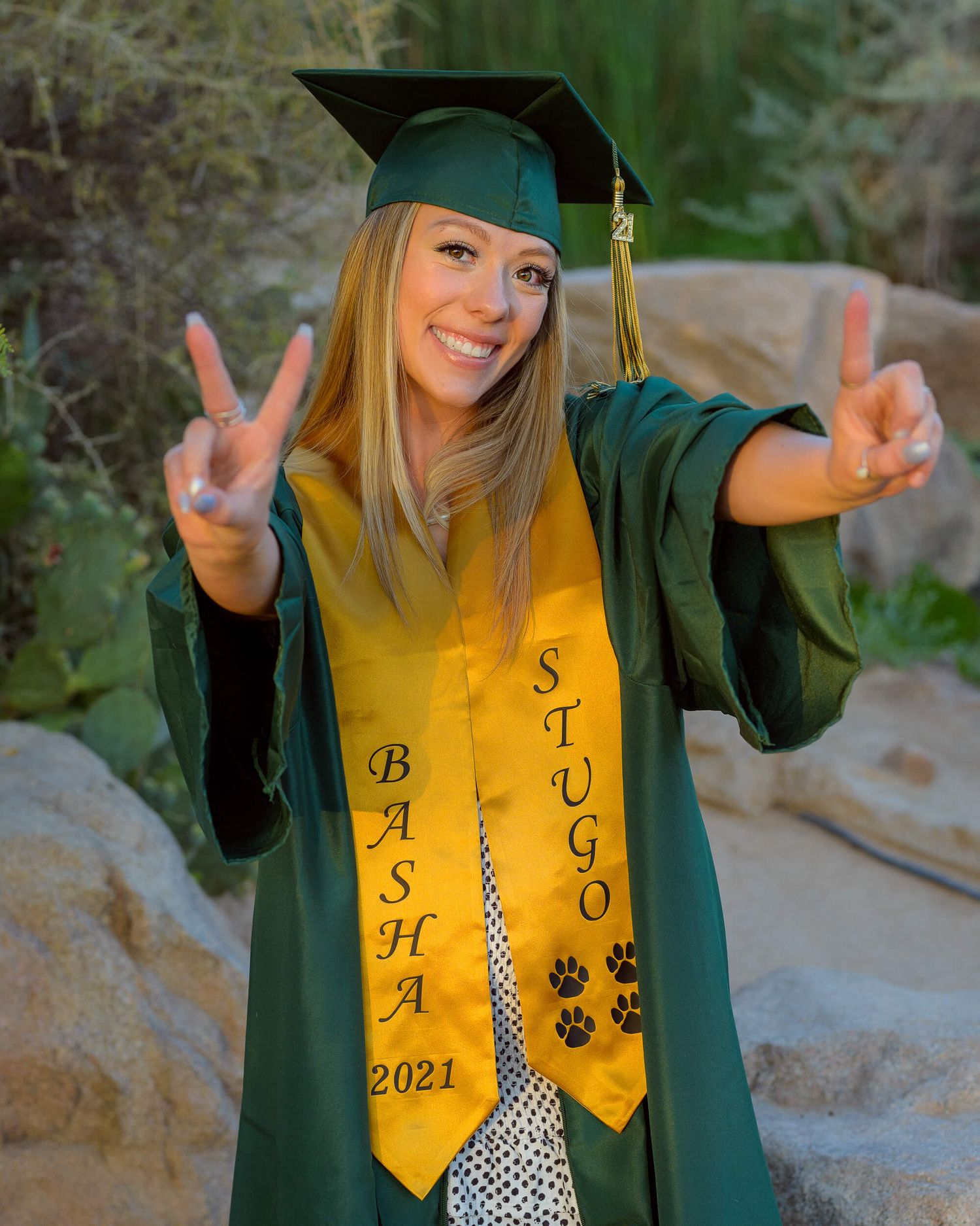 Smiling graduate in green cap and gown with gold BASHA 2021 STUGO stole making peace signs outdoors.