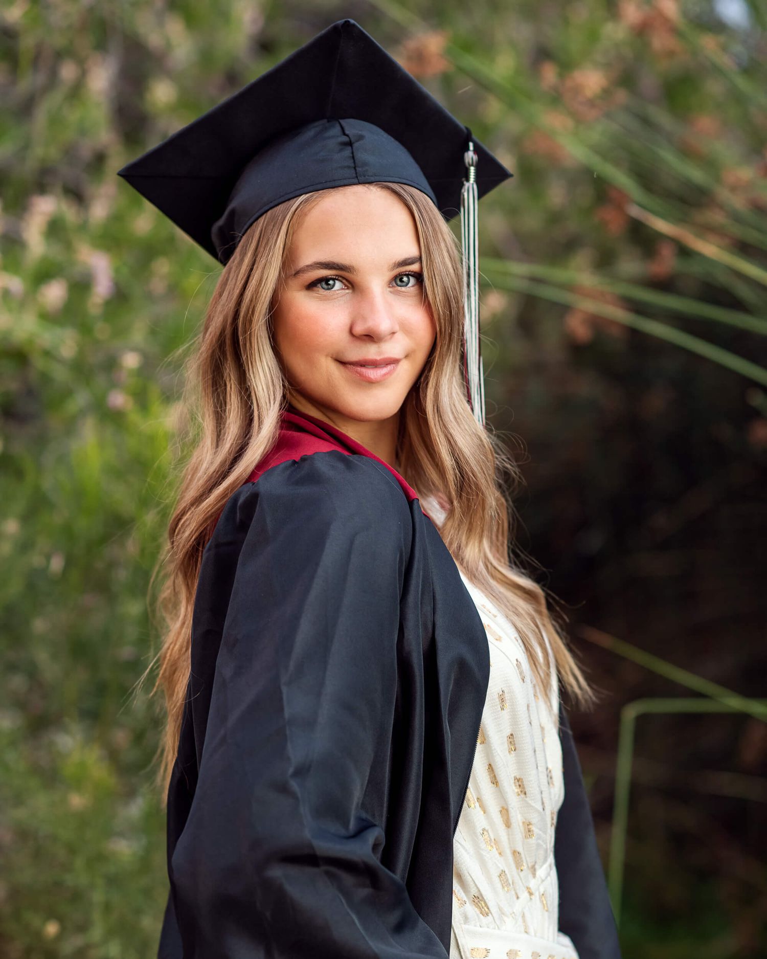 Smiling graduate in black cap and gown with red collar poses outdoors among green foliage for graduation portrait.