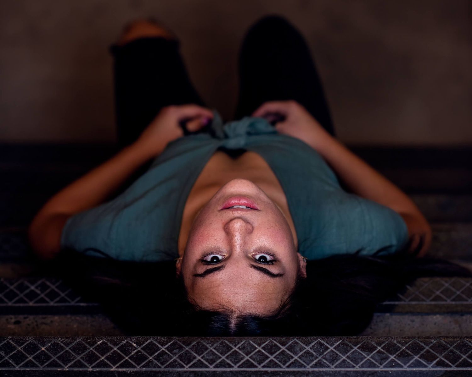 Woman in teal dress lying on stairs, looking up at camera in dramatic low-light photography.