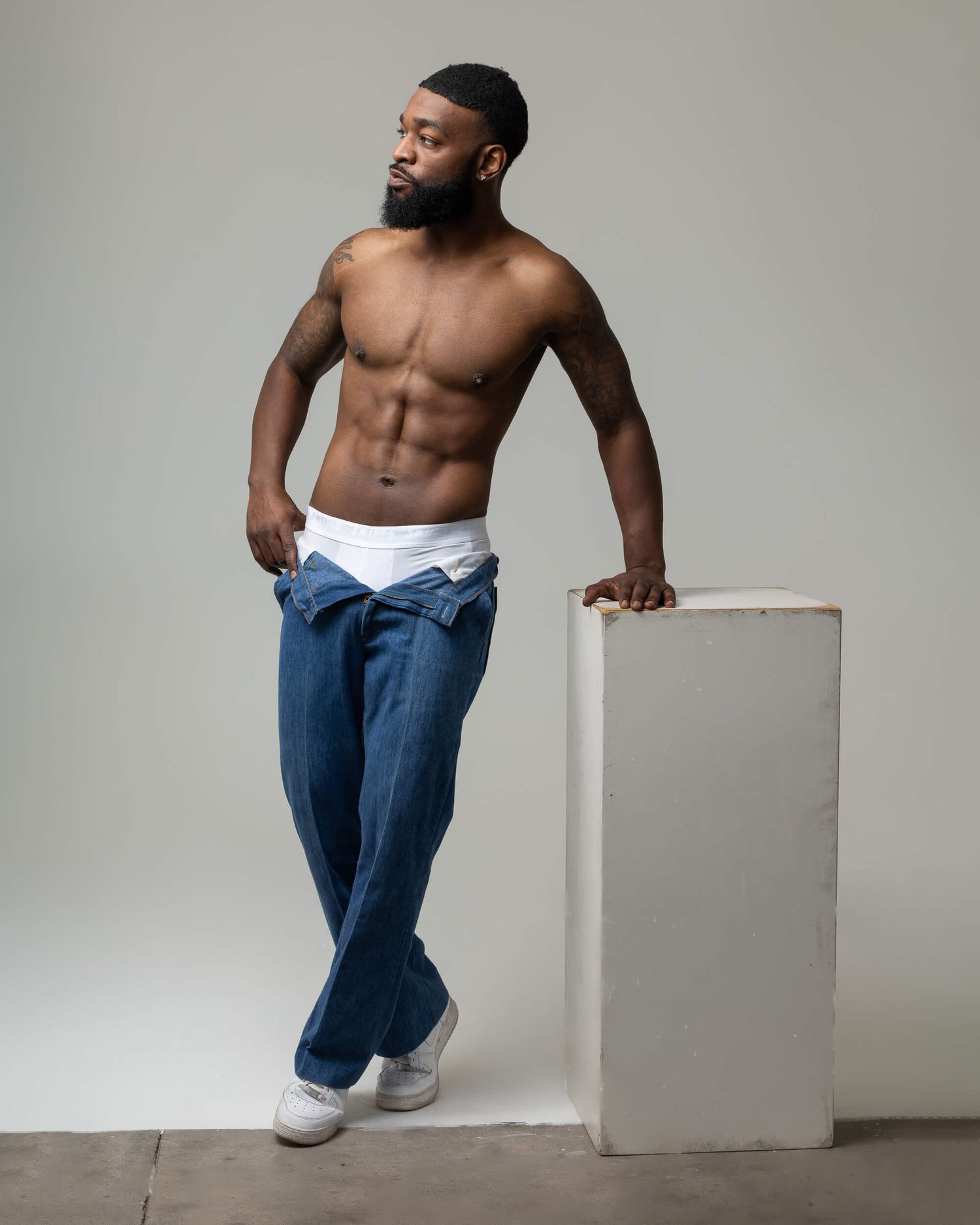 Fit shirtless Black man with beard leans on white block wearing low-rise jeans and white sneakers in studio.