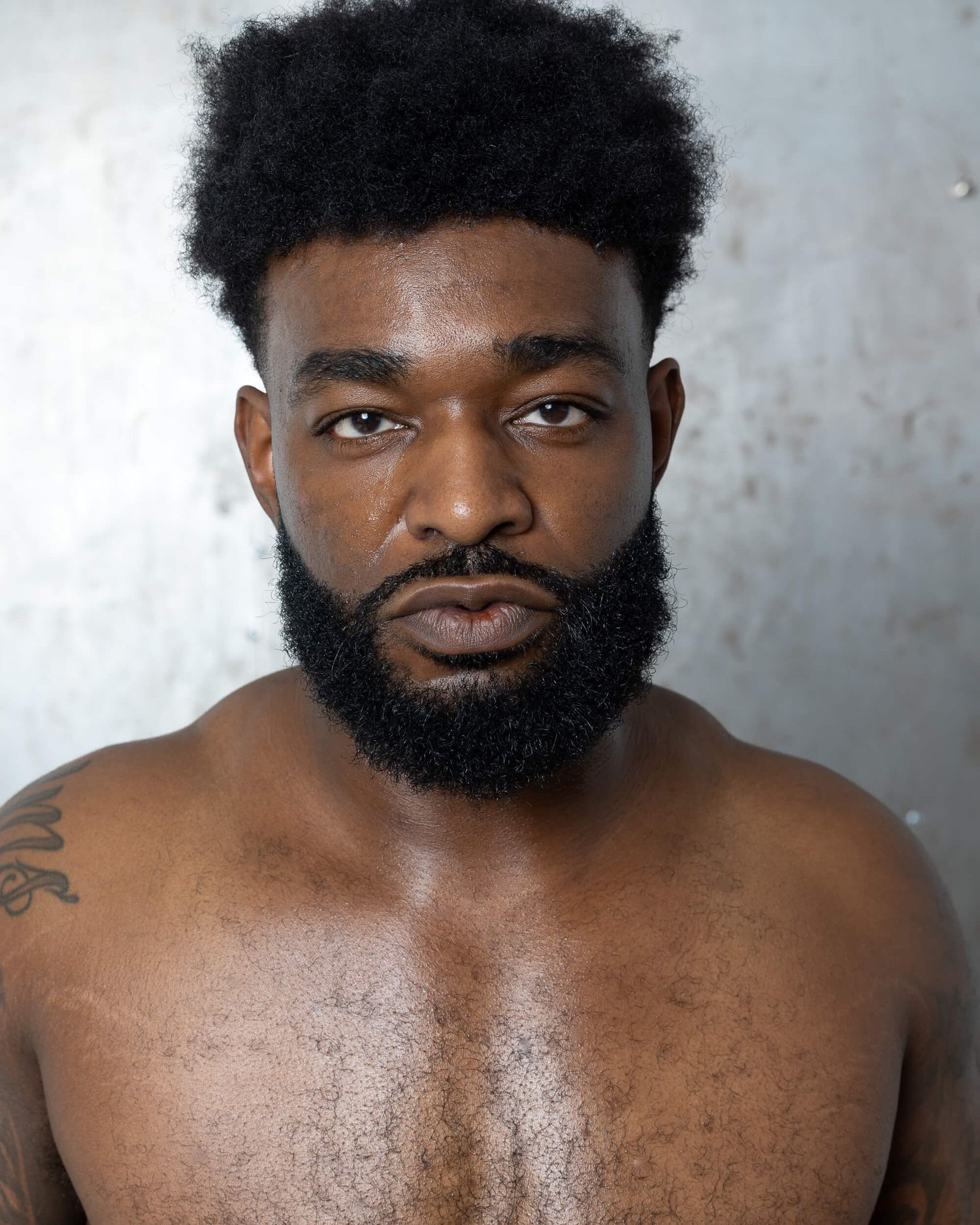 Shirtless Black man with natural afro hair and full beard poses confidently against a gray background.