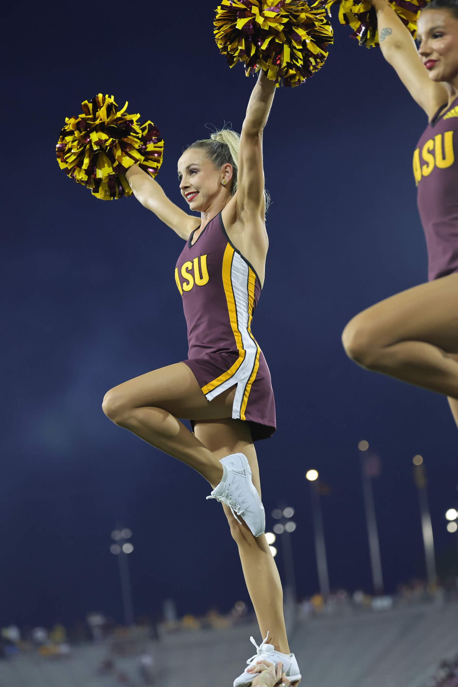 ASU cheerleader in maroon uniform raises gold pom-poms while performing at night game.