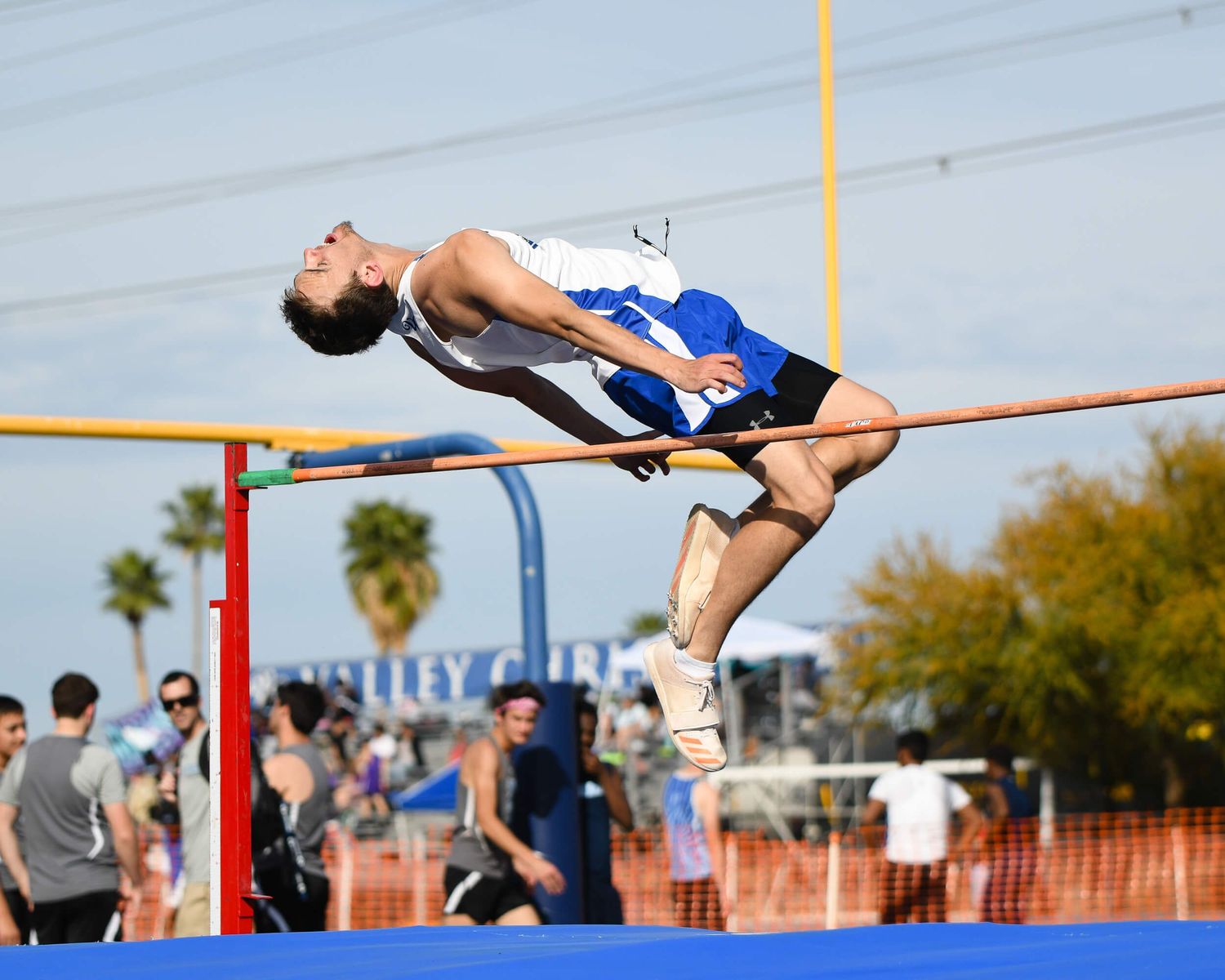 Male high jumper in blue and white uniform clears the bar using the Fosbury Flop at an outdoor track meet.
