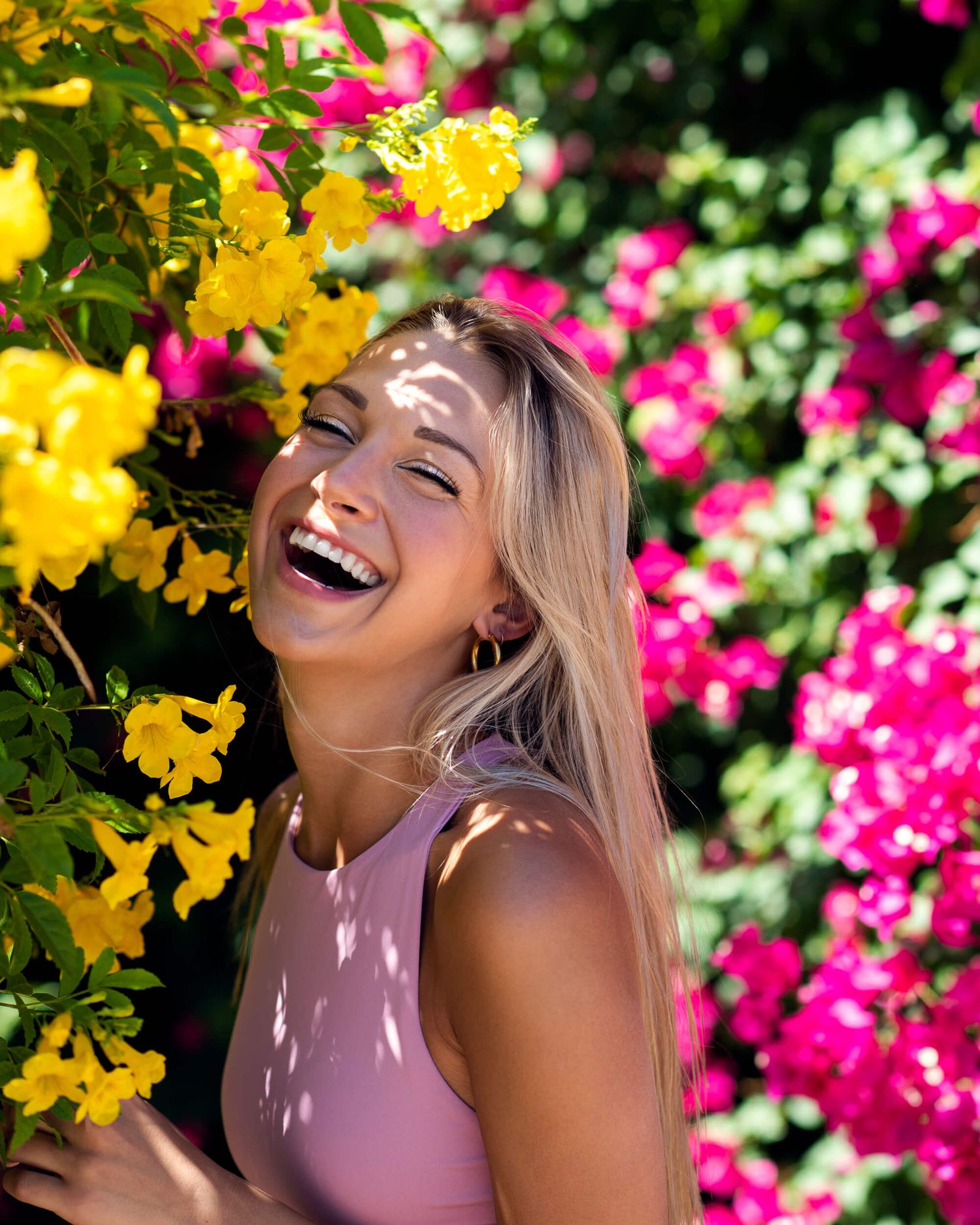Laughing blonde woman in pink top surrounded by vibrant yellow and pink blooming flowers.