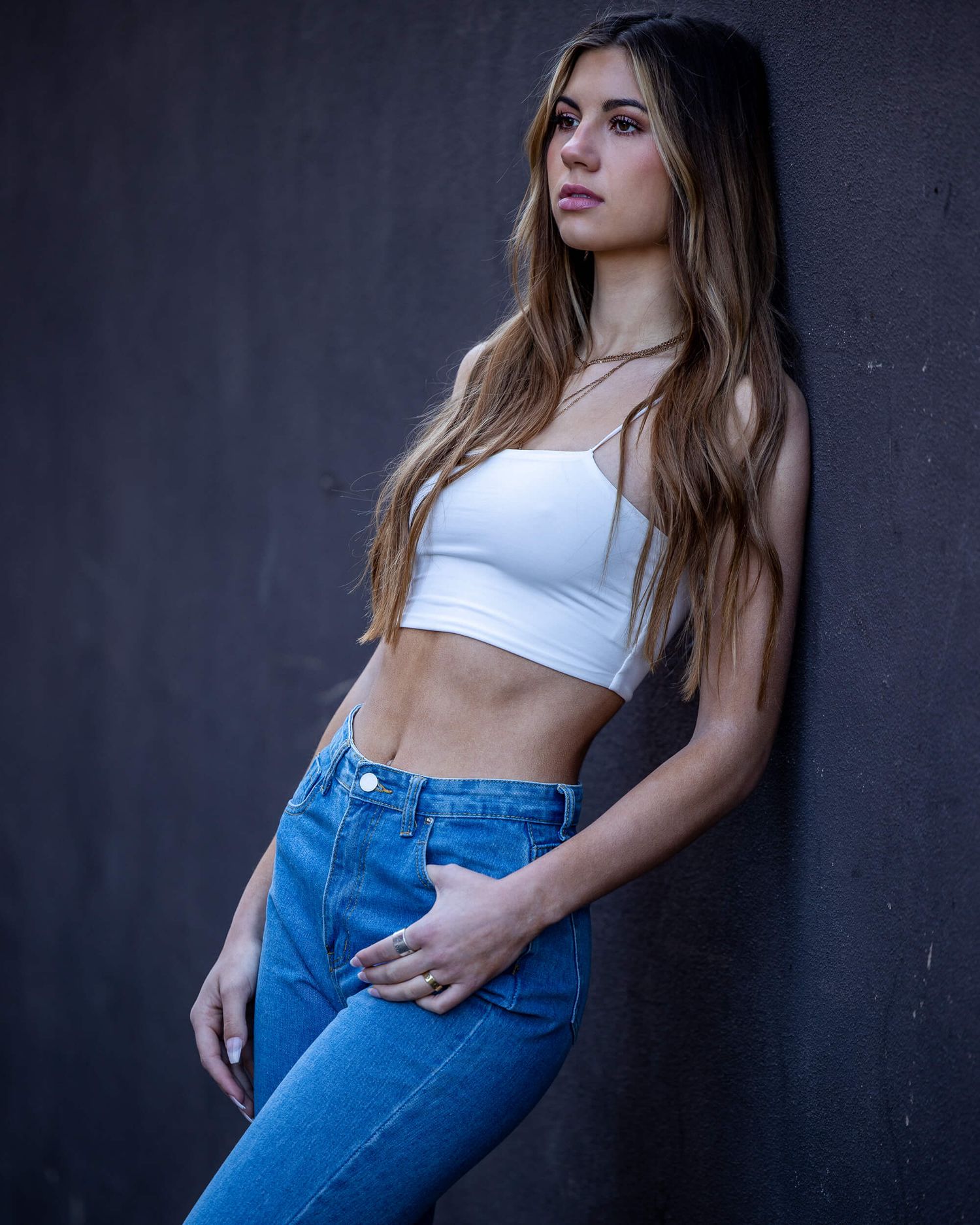 Young woman in white crop top and blue jeans leaning against a dark gray wall.