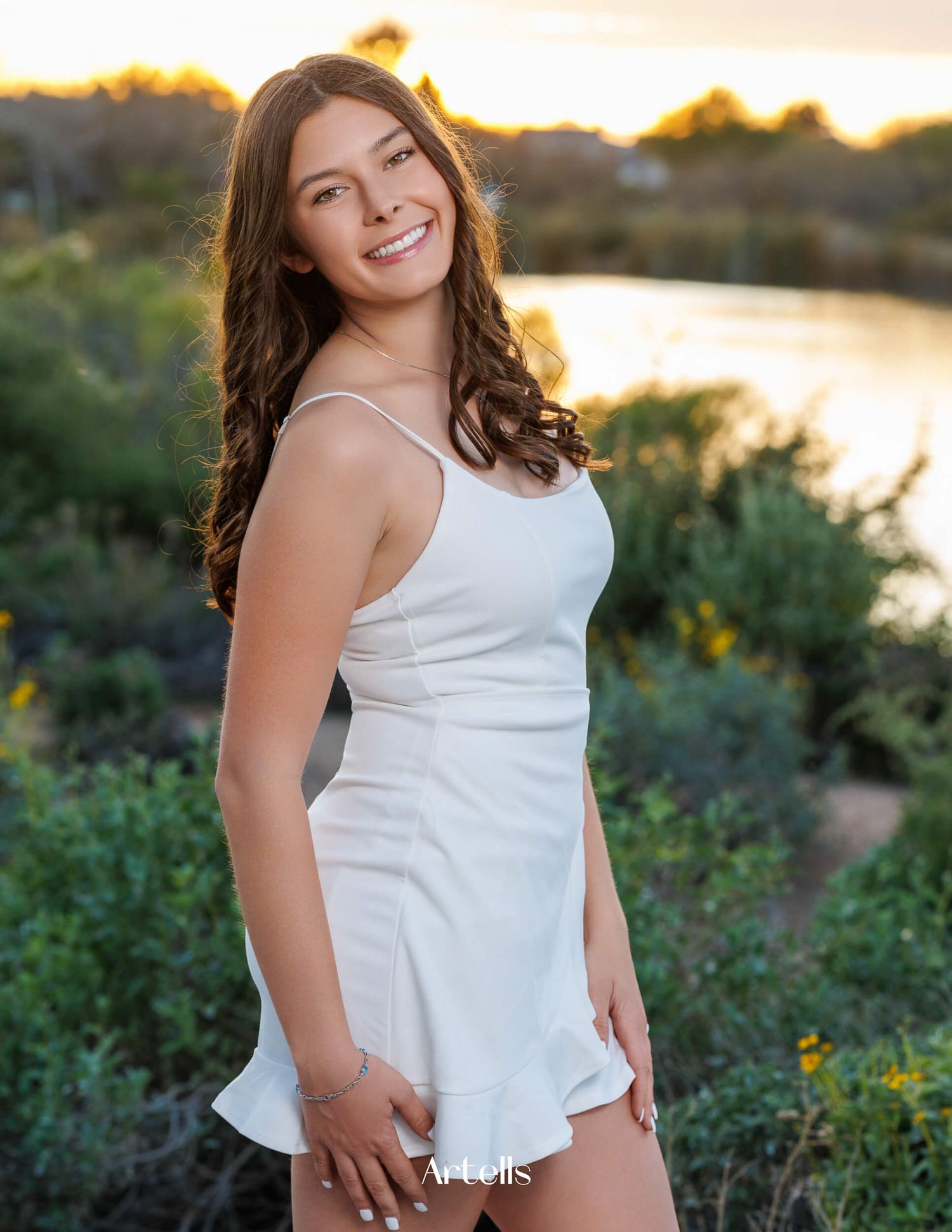 Smiling young woman in white sundress poses outdoors at golden hour by a scenic waterway.
