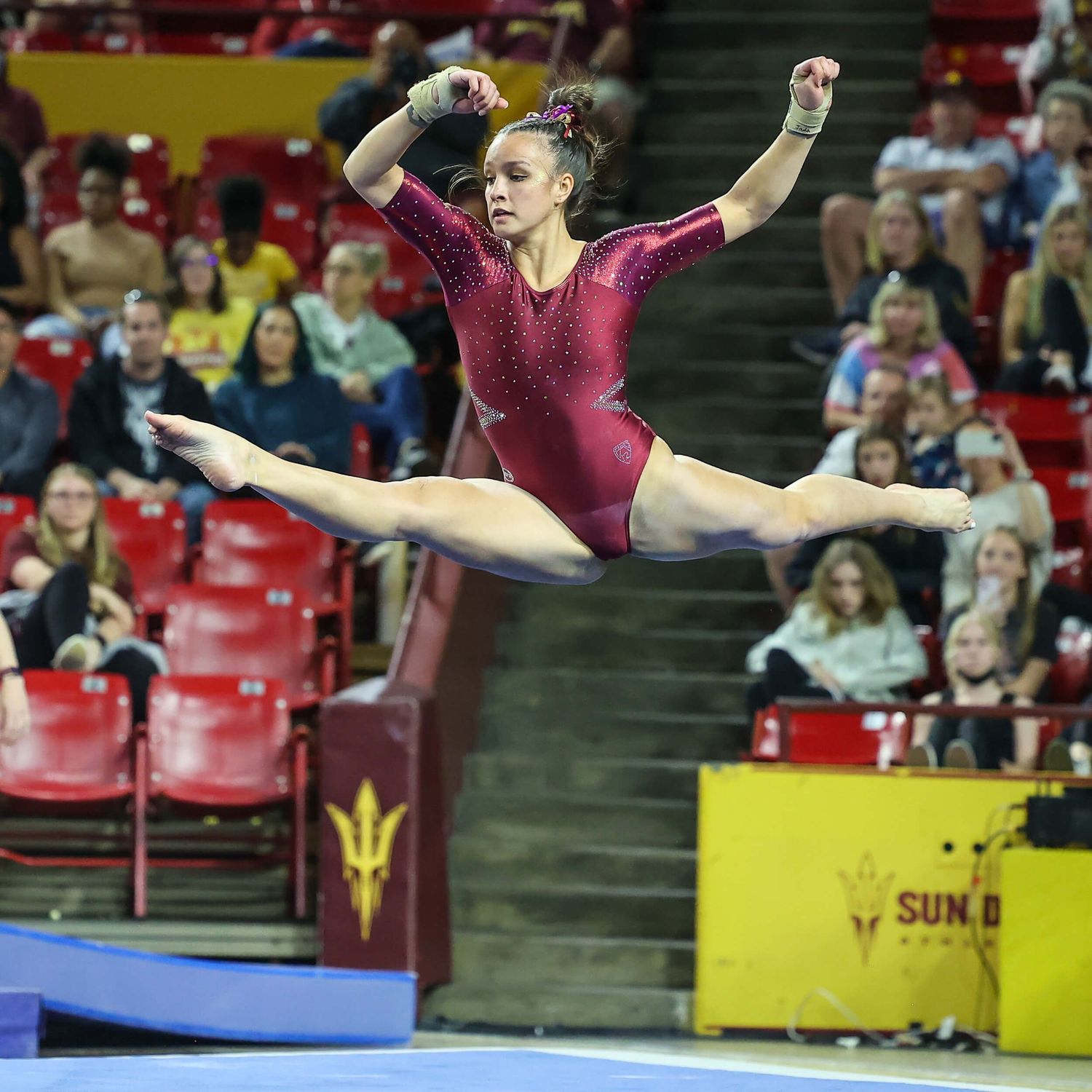 Female gymnast in red leotard performs a split leap during a college gymnastics competition at an indoor arena.
