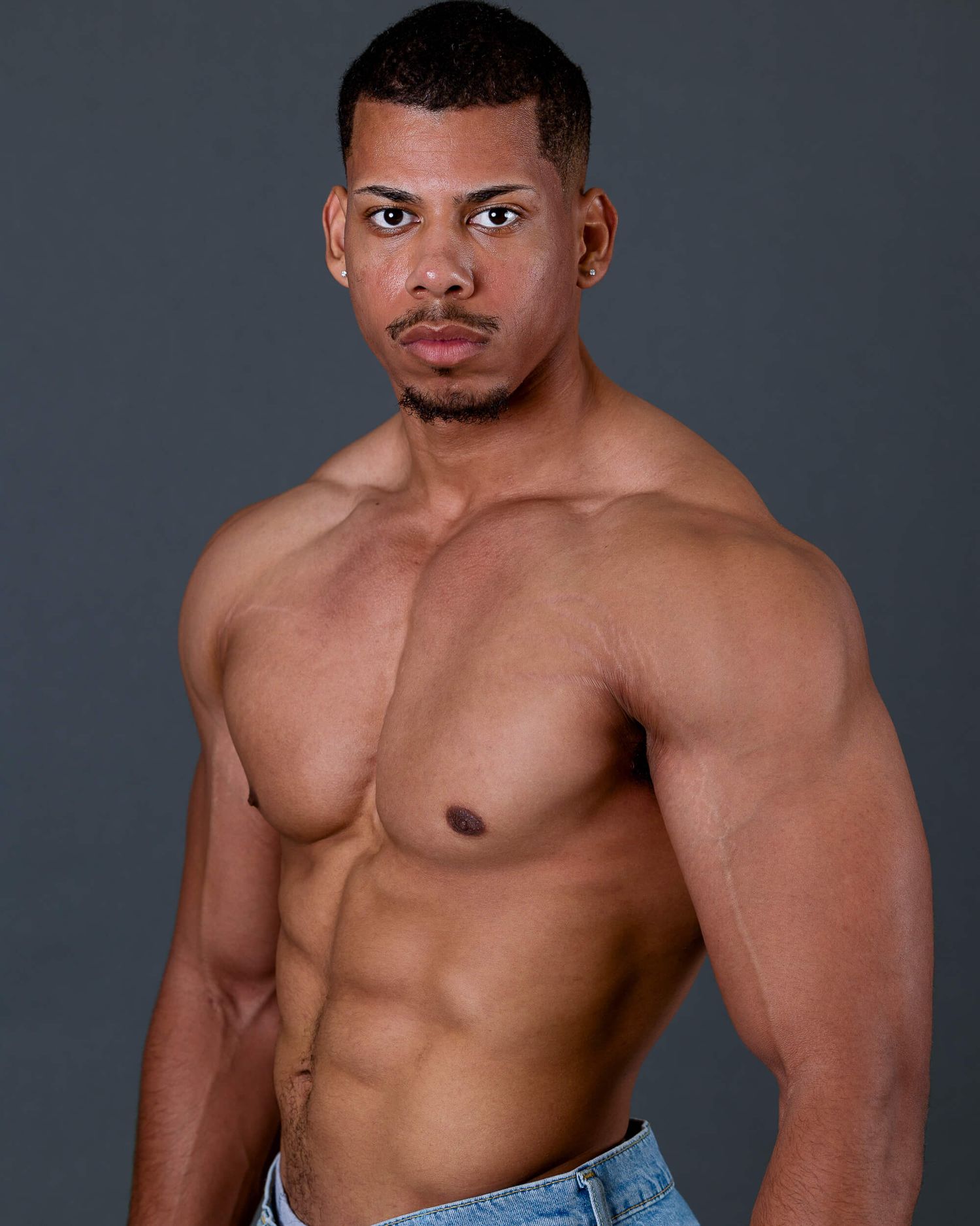 Muscular shirtless man with defined abs posing in open blue jeans against dark gray background.