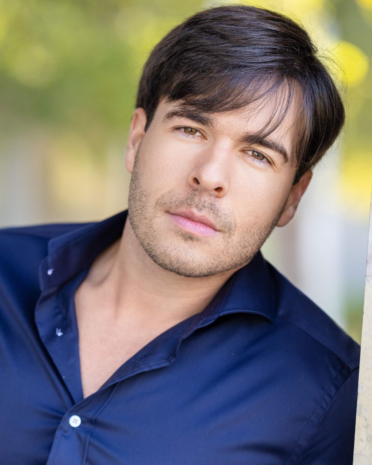 Handsome dark-haired man with blue eyes poses outdoors in a navy blue button-up shirt.