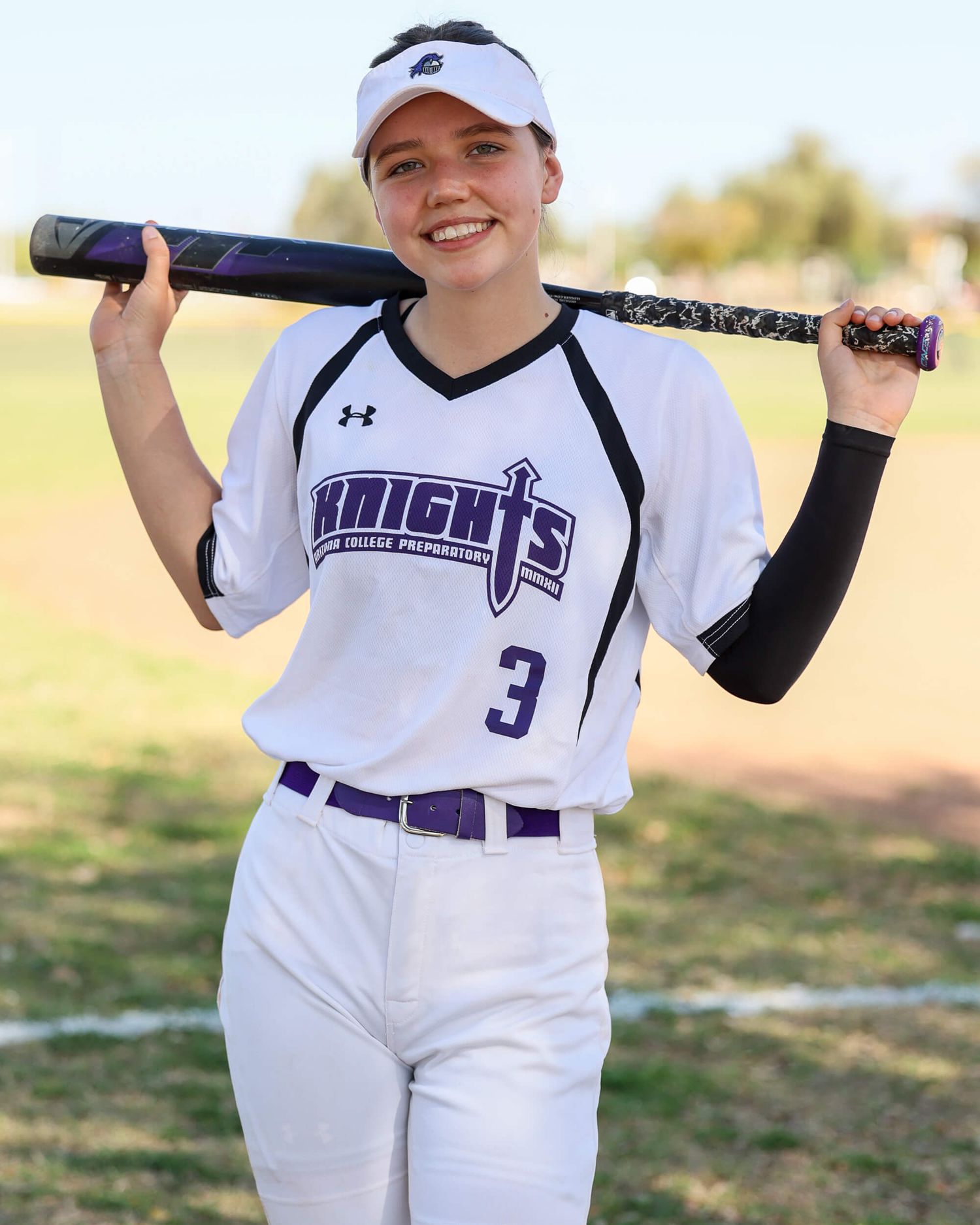 A smiling female softball player in a Knights uniform holds a bat over her shoulder outdoors.