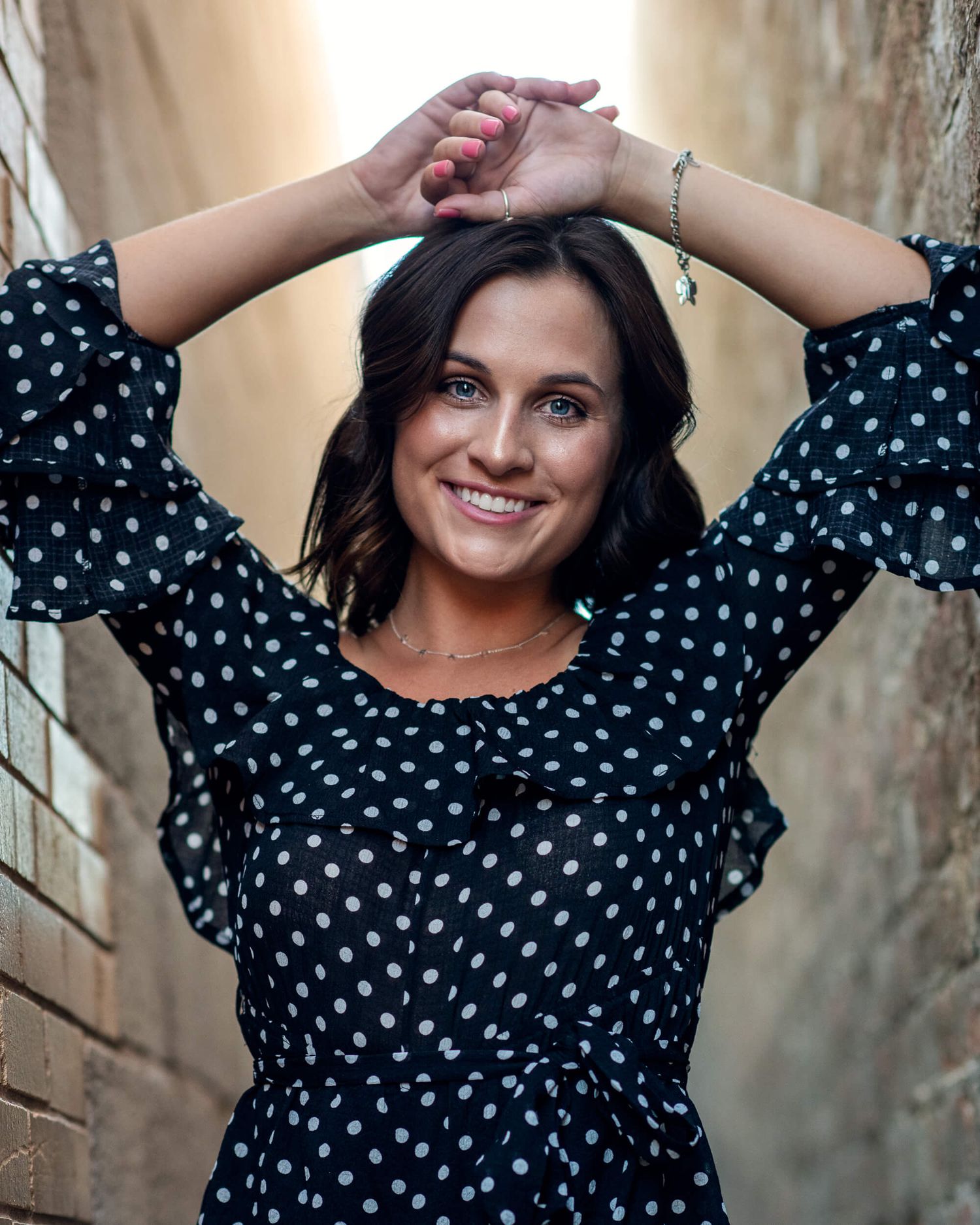 Smiling woman in polka dot dress posing in sunlit brick alley.