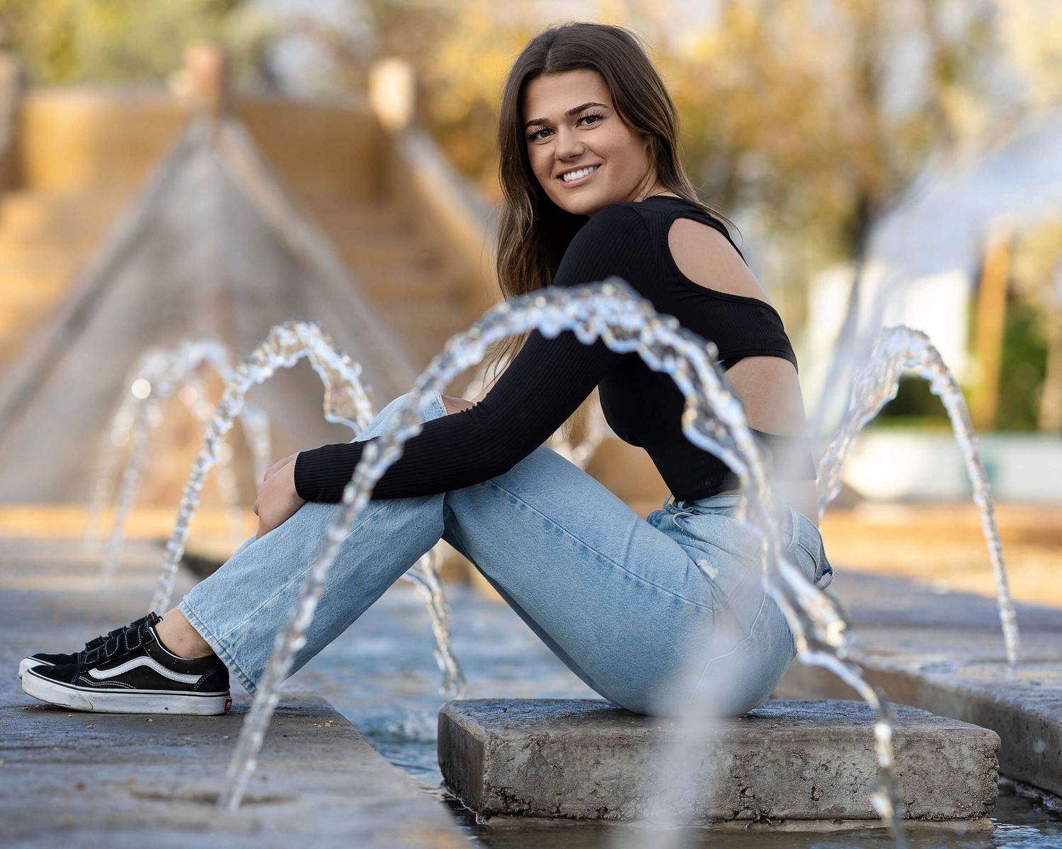 Smiling young woman in jeans sitting by fountain outdoors.