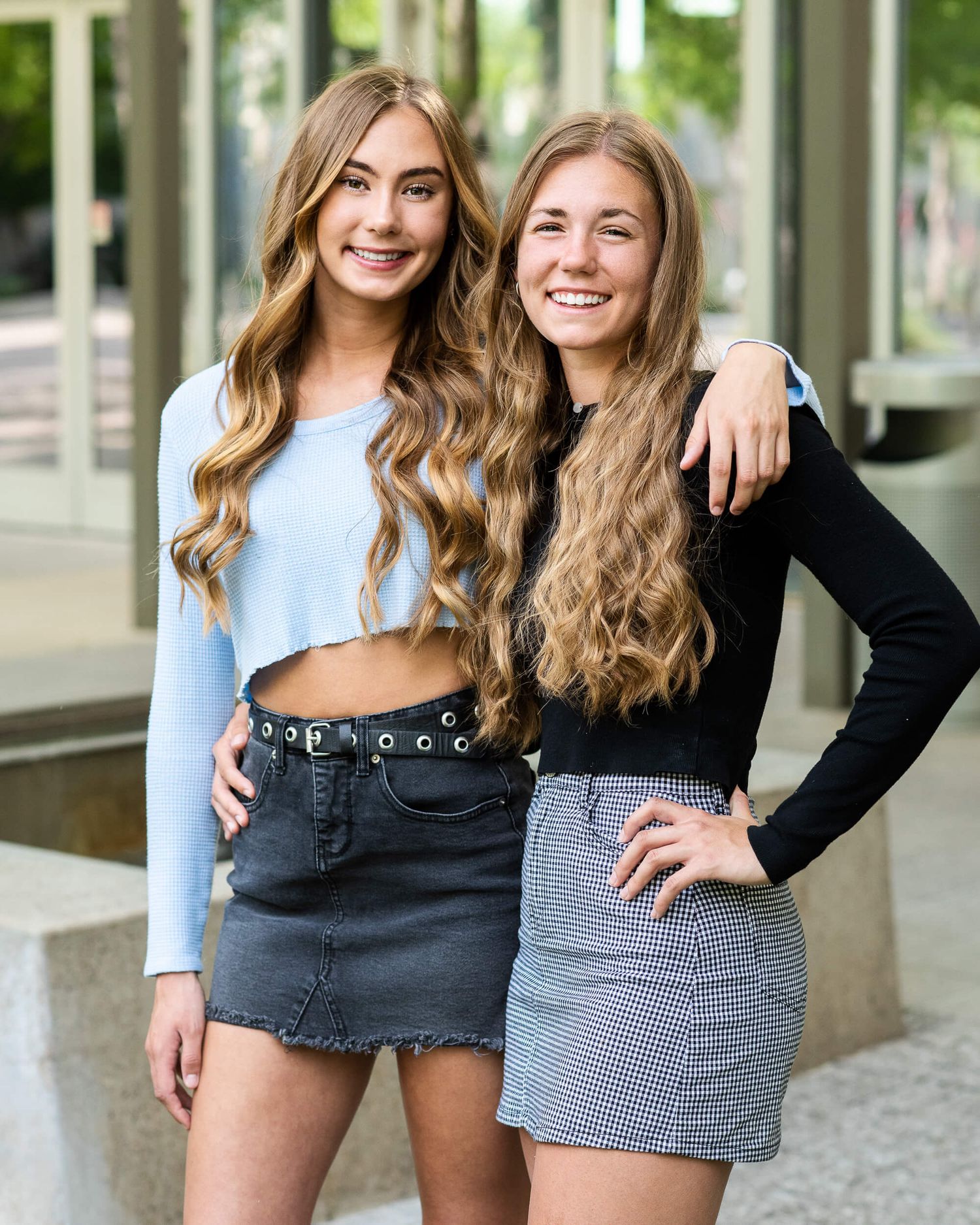 Two smiling young women in crop tops and mini skirts posing together outdoors.