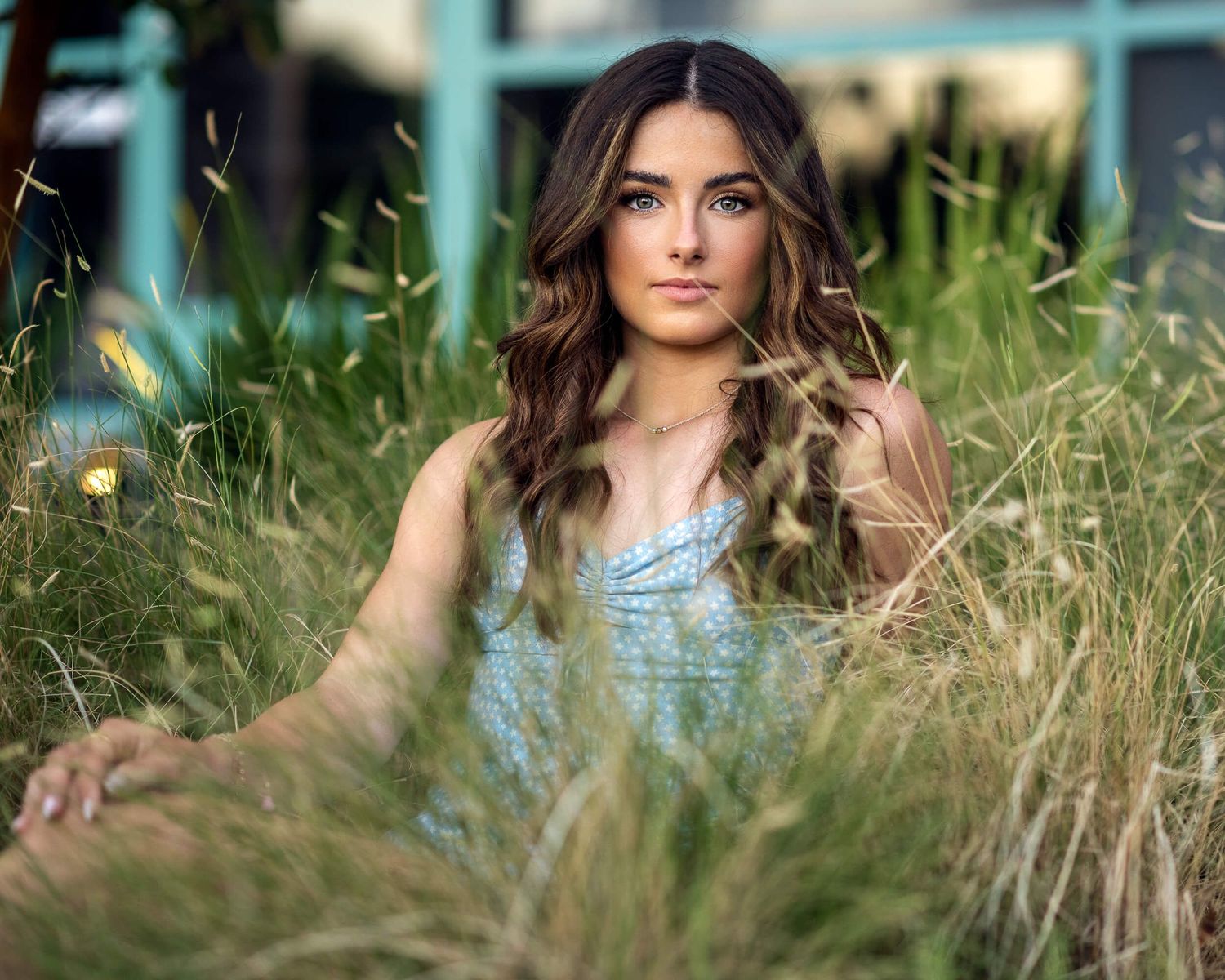 Young woman with long wavy brunette hair in a light blue dress posing gracefully among tall green grass outdoors.