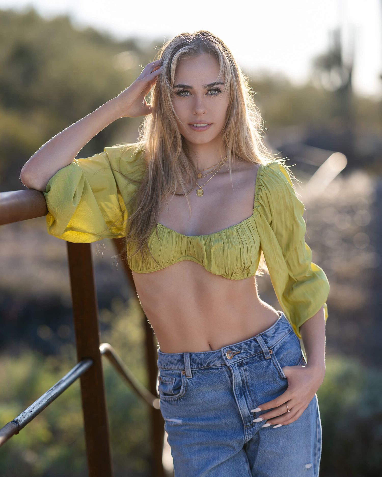 Blonde woman in yellow cropped puff-sleeve top and jeans posing by a railing in a sunny outdoor setting.