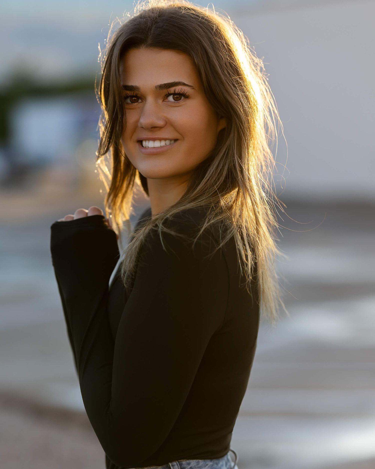 Young woman with long hair smiling over shoulder in golden sunlight, wearing black top outdoors.
