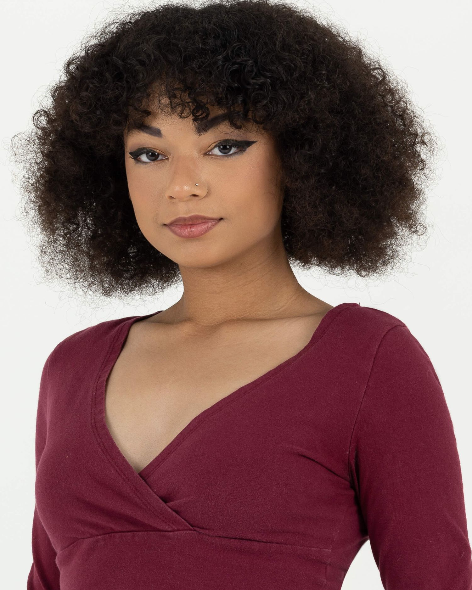 Young woman with natural curly hair wearing a burgundy wrap top poses against white background.